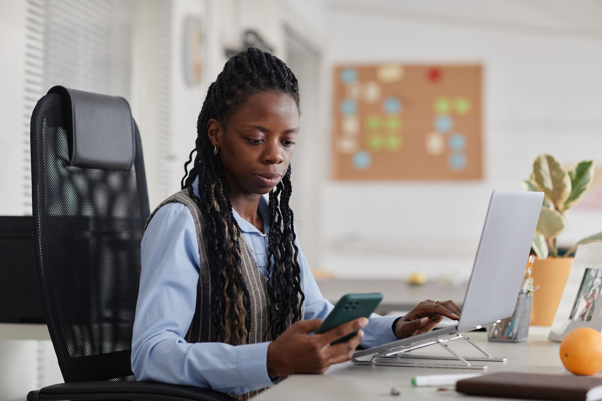 A woman is sitting at a desk using a laptop and a cell phone.