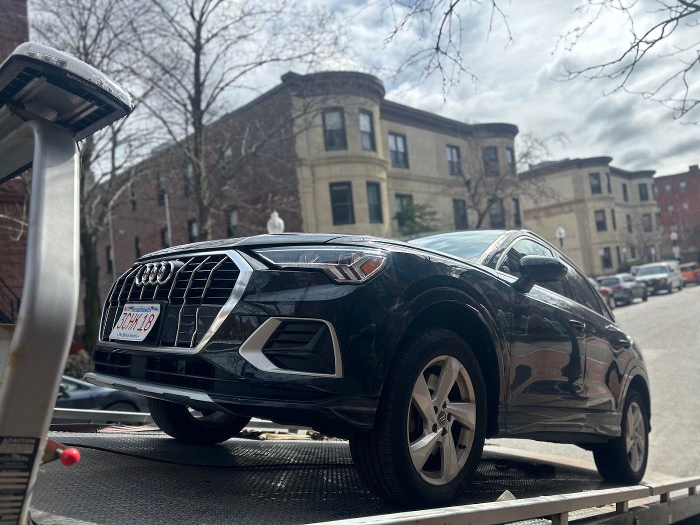 Black Audi Q3 SUV on a tow truck, parked on a city street with brick buildings.