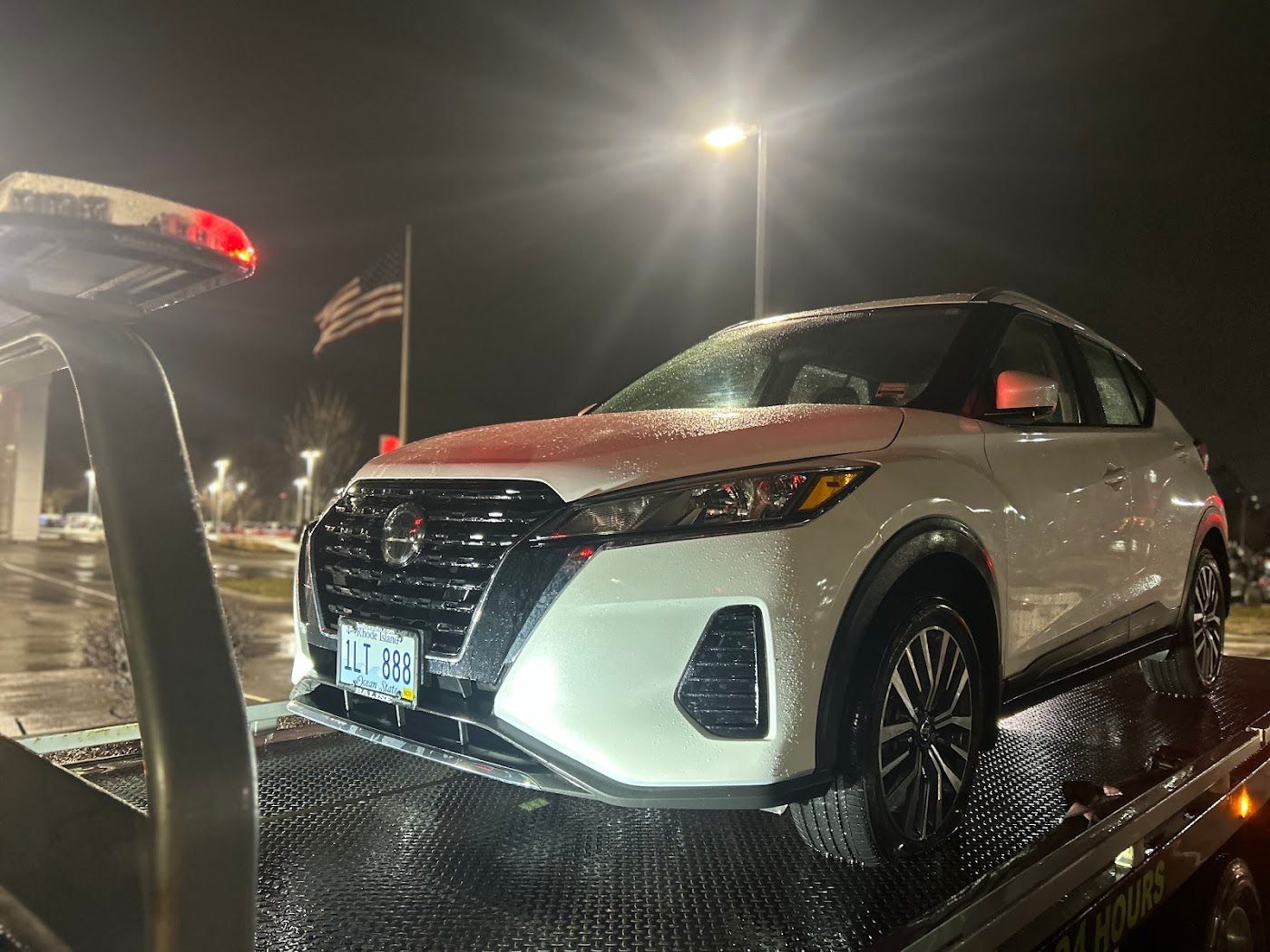 White SUV on a tow truck at night, with wet pavement and bright overhead light.