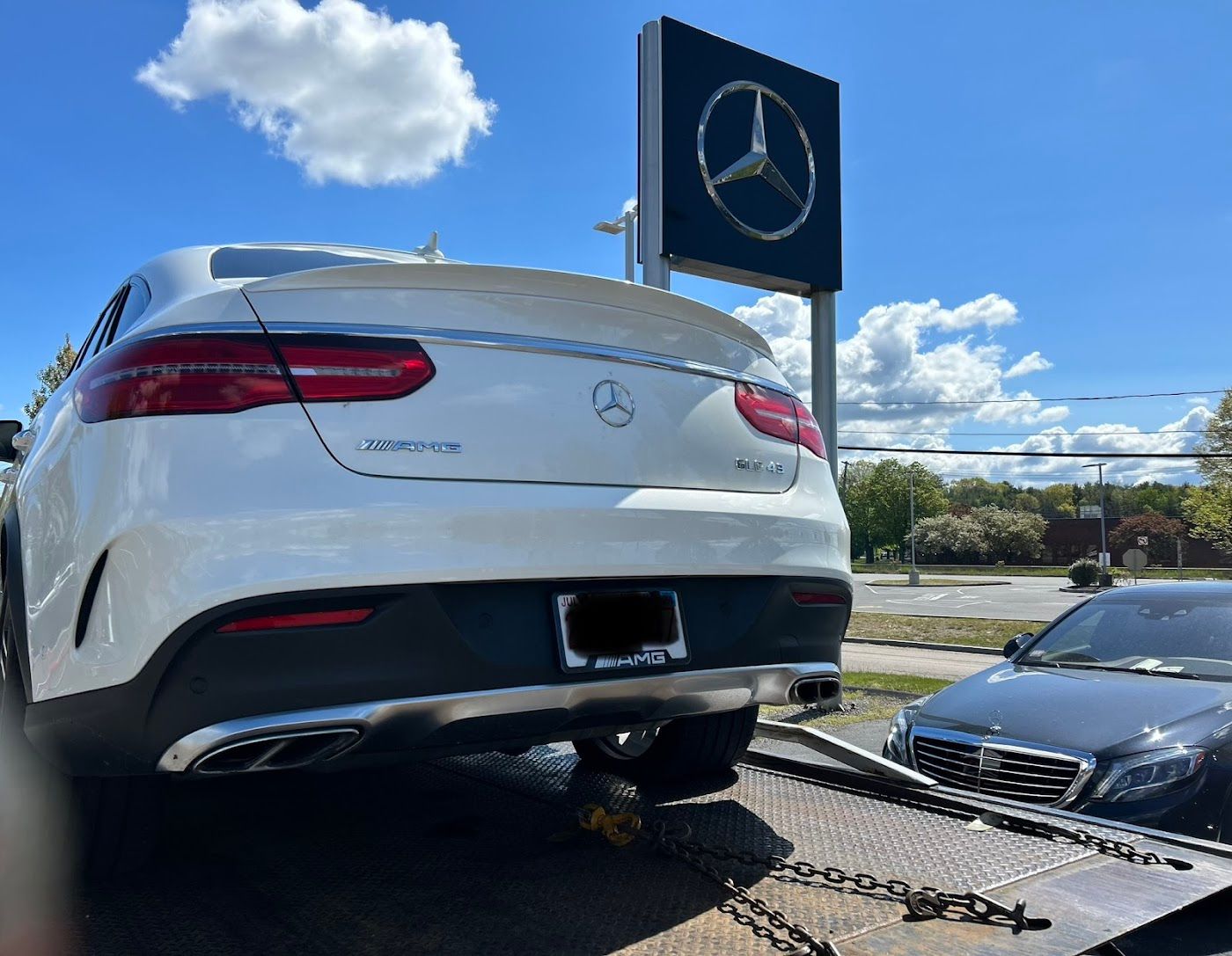 White Mercedes-Benz SUV on a tow truck, parked near a Mercedes-Benz dealership sign, sunny day.