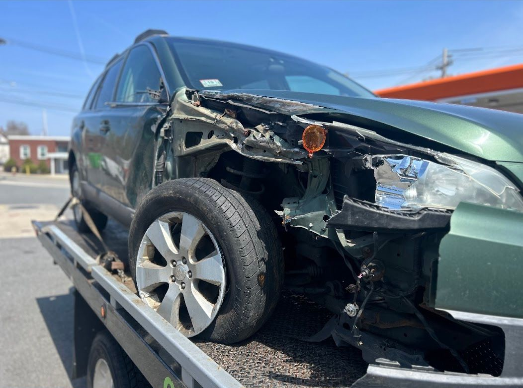 Green SUV with front-end damage, being towed on a flatbed truck on a sunny day.