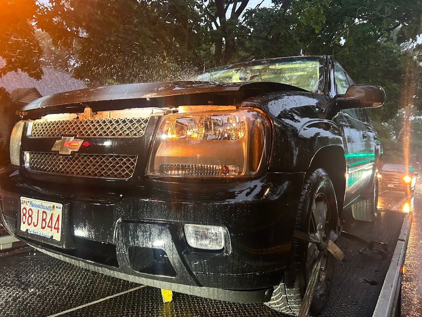 Black Chevrolet Trailblazer on a tow truck, damaged windshield, front end, headlights on, water spray.