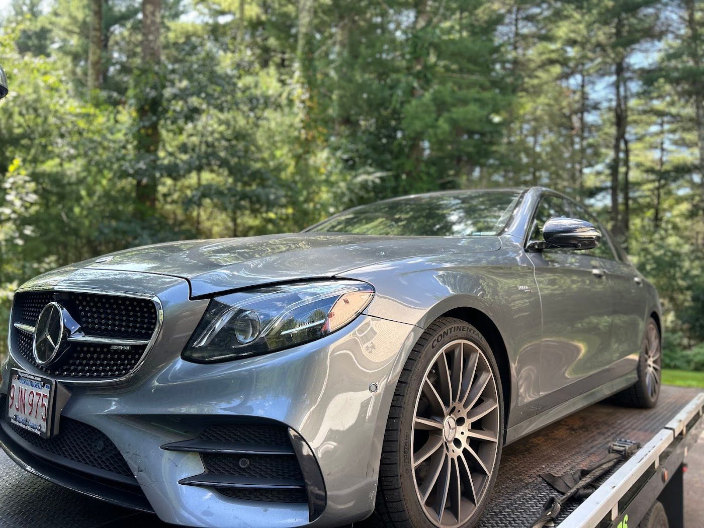 Silver Mercedes-Benz sedan on a tow truck, outdoors with a green, leafy background.