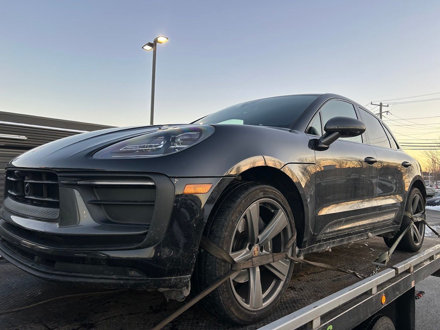 Black Porsche SUV on a tow truck, secured with straps. Snowy ground, outdoor setting.