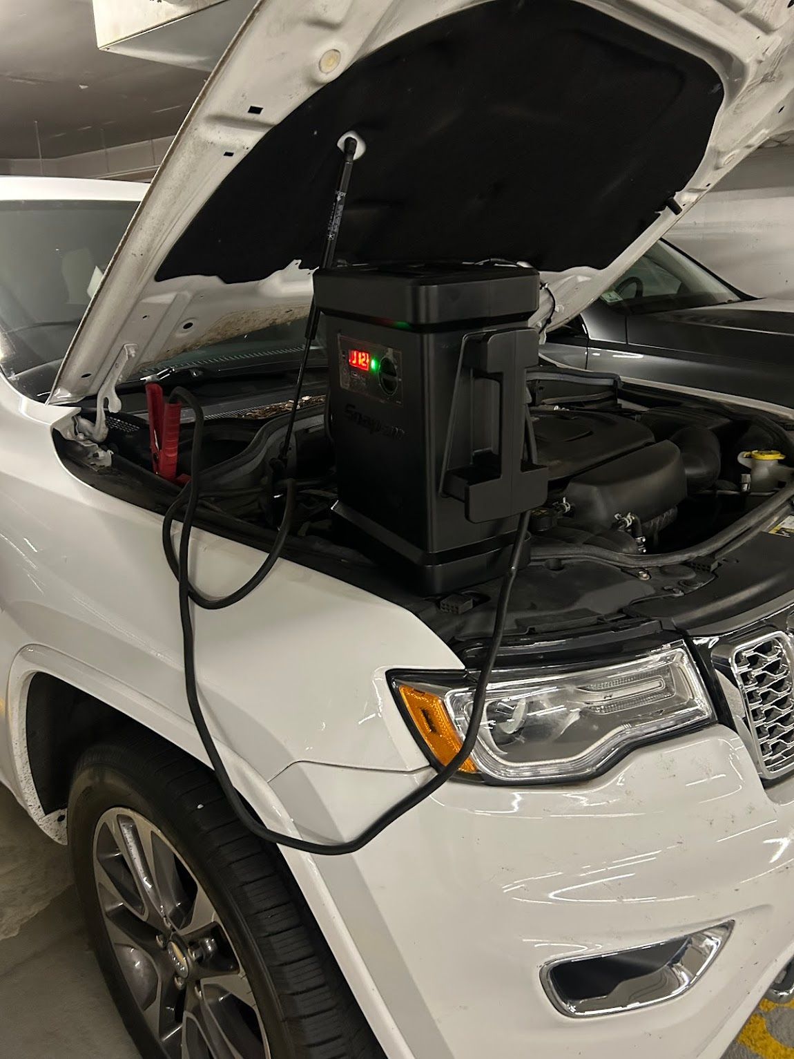 A white SUV with its hood up, jumper cables attached to a black device in the engine.