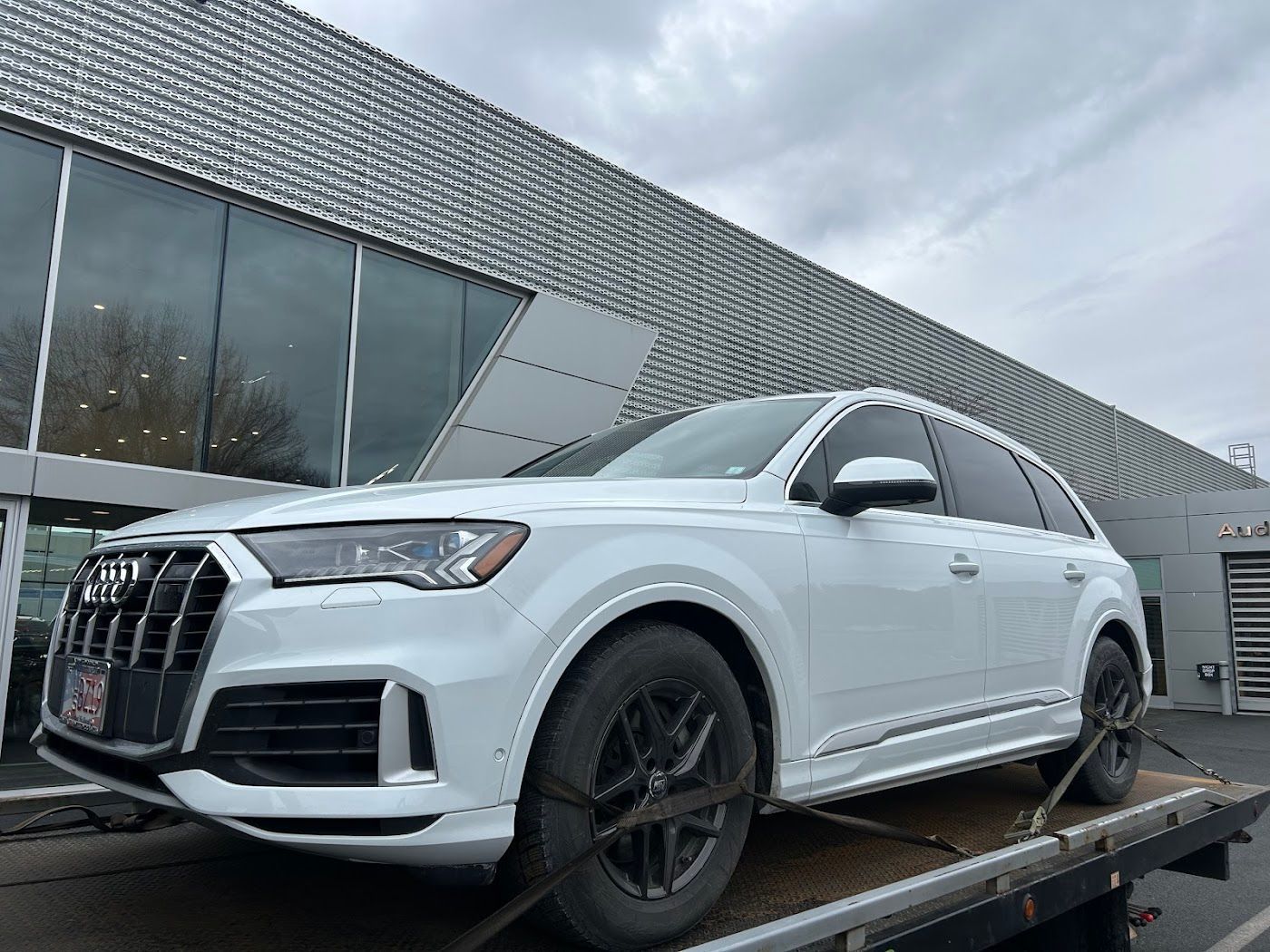 White Audi Q7 SUV on a flatbed tow truck in front of a modern building with large windows.
