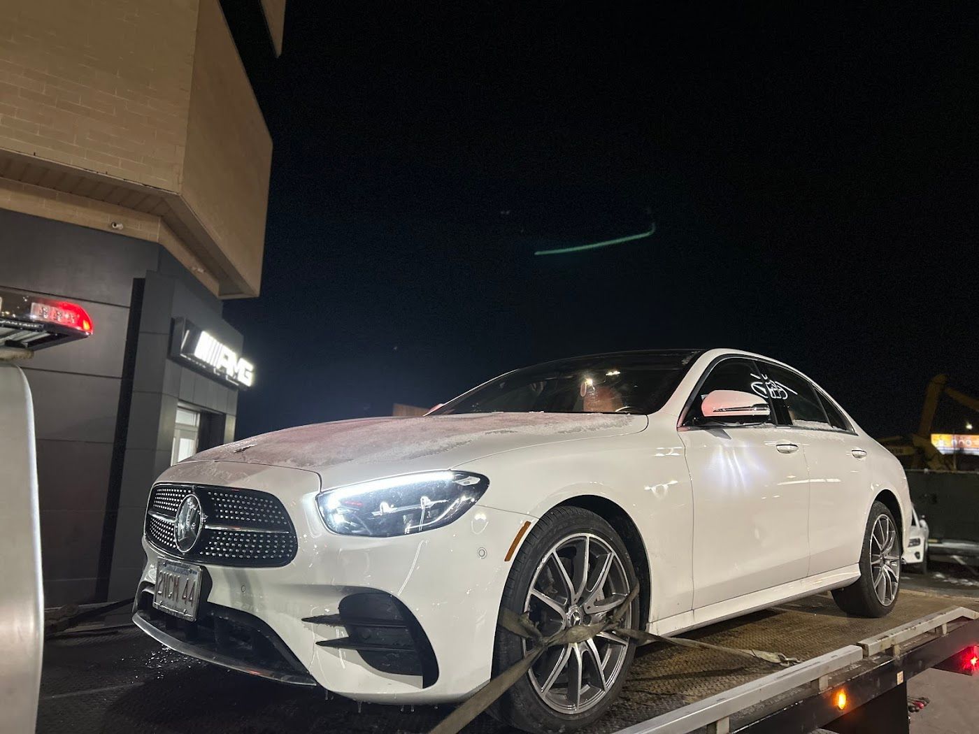 White Mercedes-Benz sedan on a tow truck at night, parked in front of a Mercedes dealership.