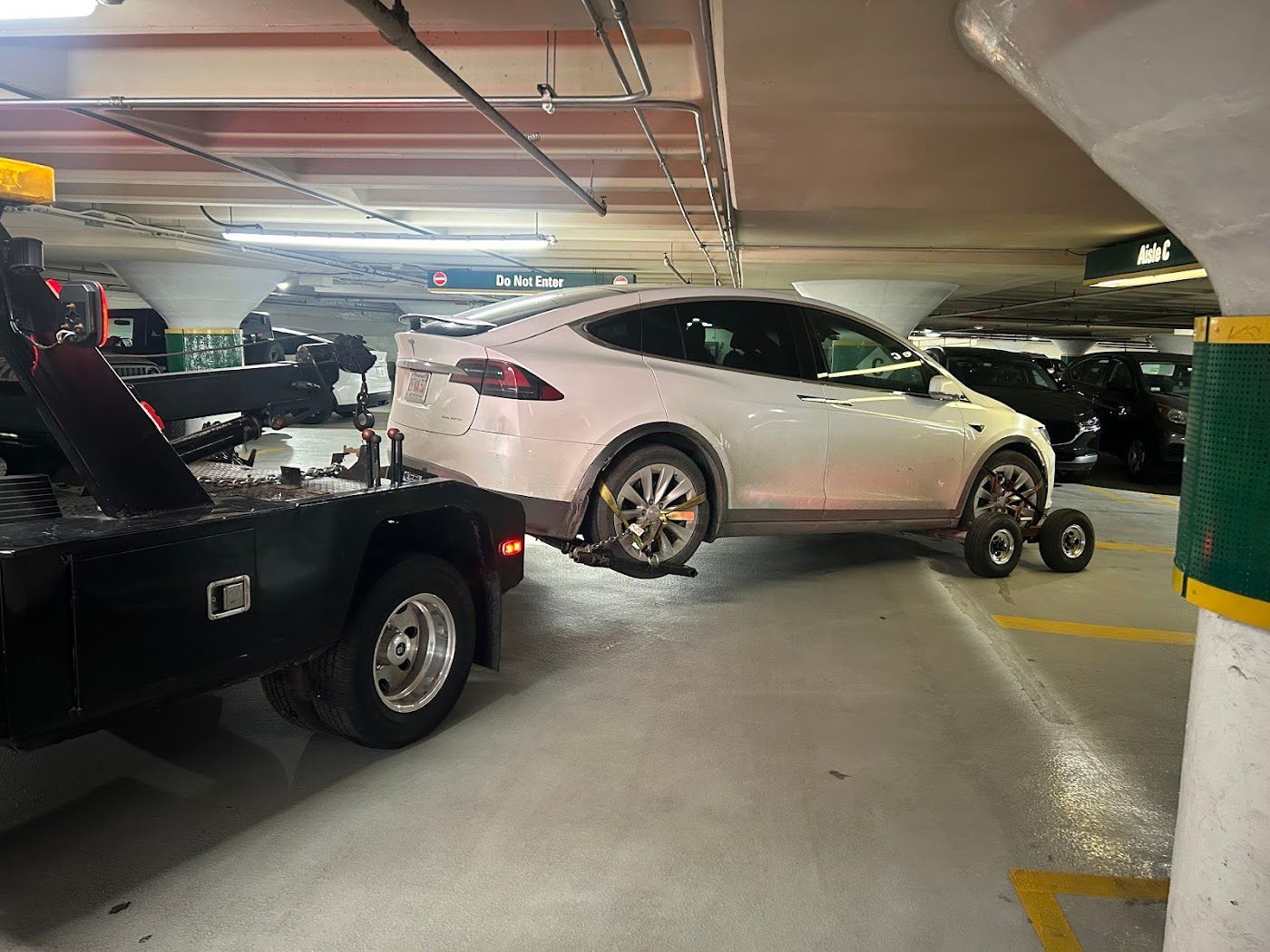 White Tesla SUV being towed in a parking garage; front wheels on dollies, connected to a tow truck.