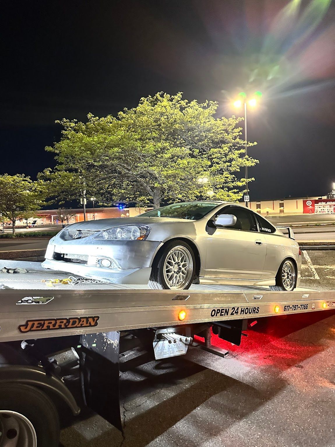 Silver car on a tow truck at night, under a tree, near a parking lot.
