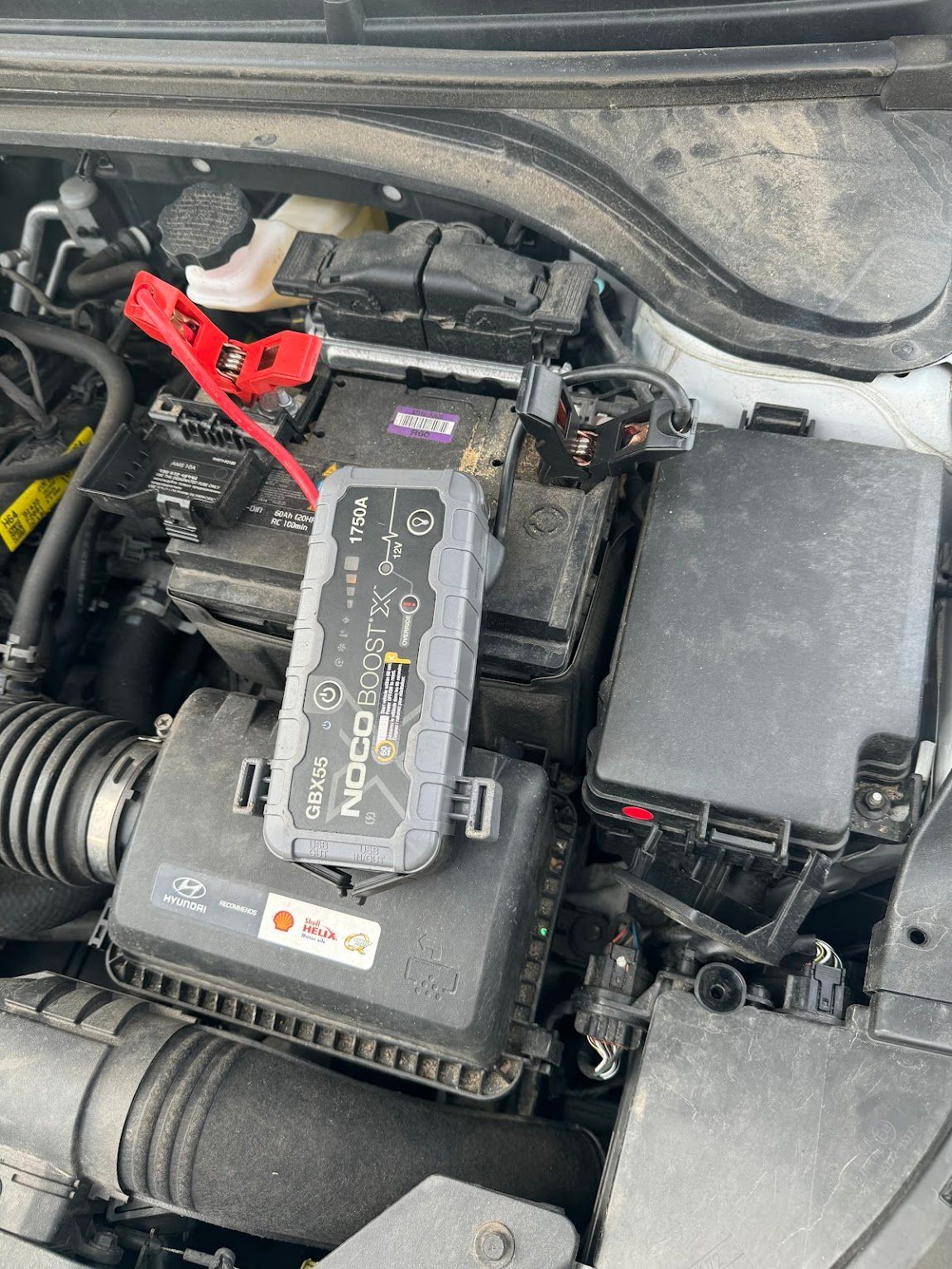 Car engine bay with a jump starter connected to the battery. Black, grey, and red cables visible.