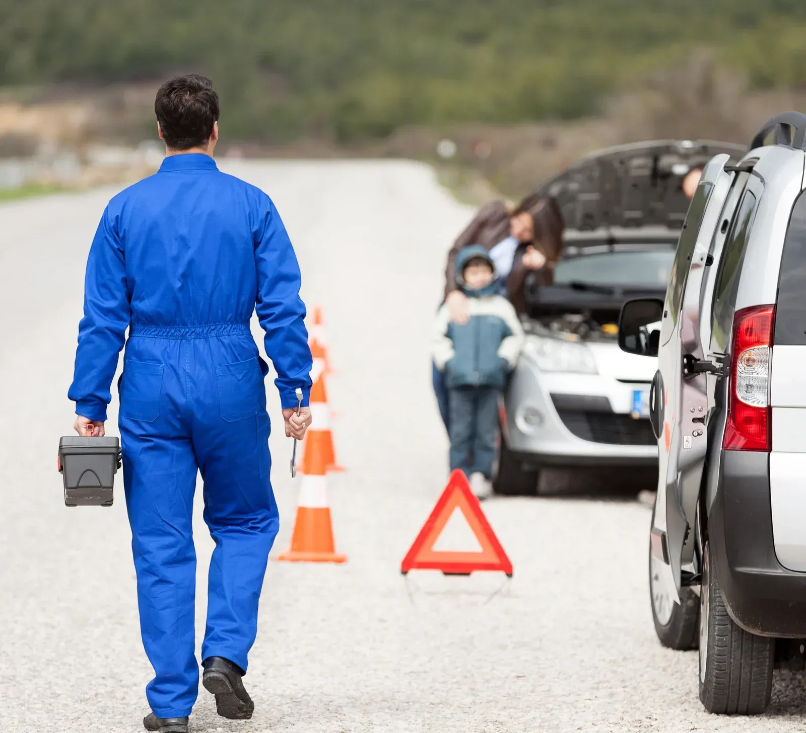 Mechanic walking toward a broken-down car on a roadside. Family stands near open hood.