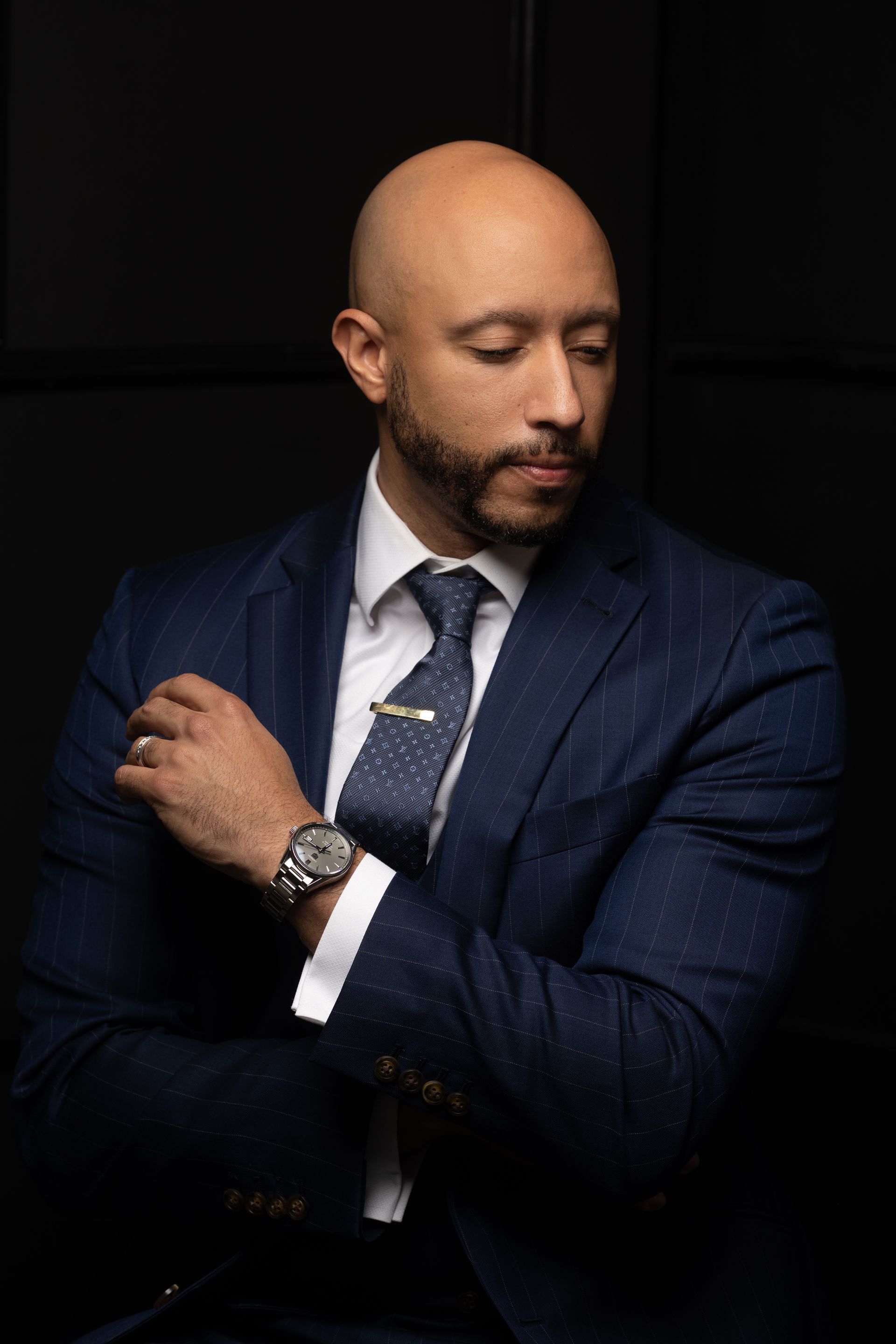 Man in a blue suit, arms crossed, looking down pensively against a dark background.