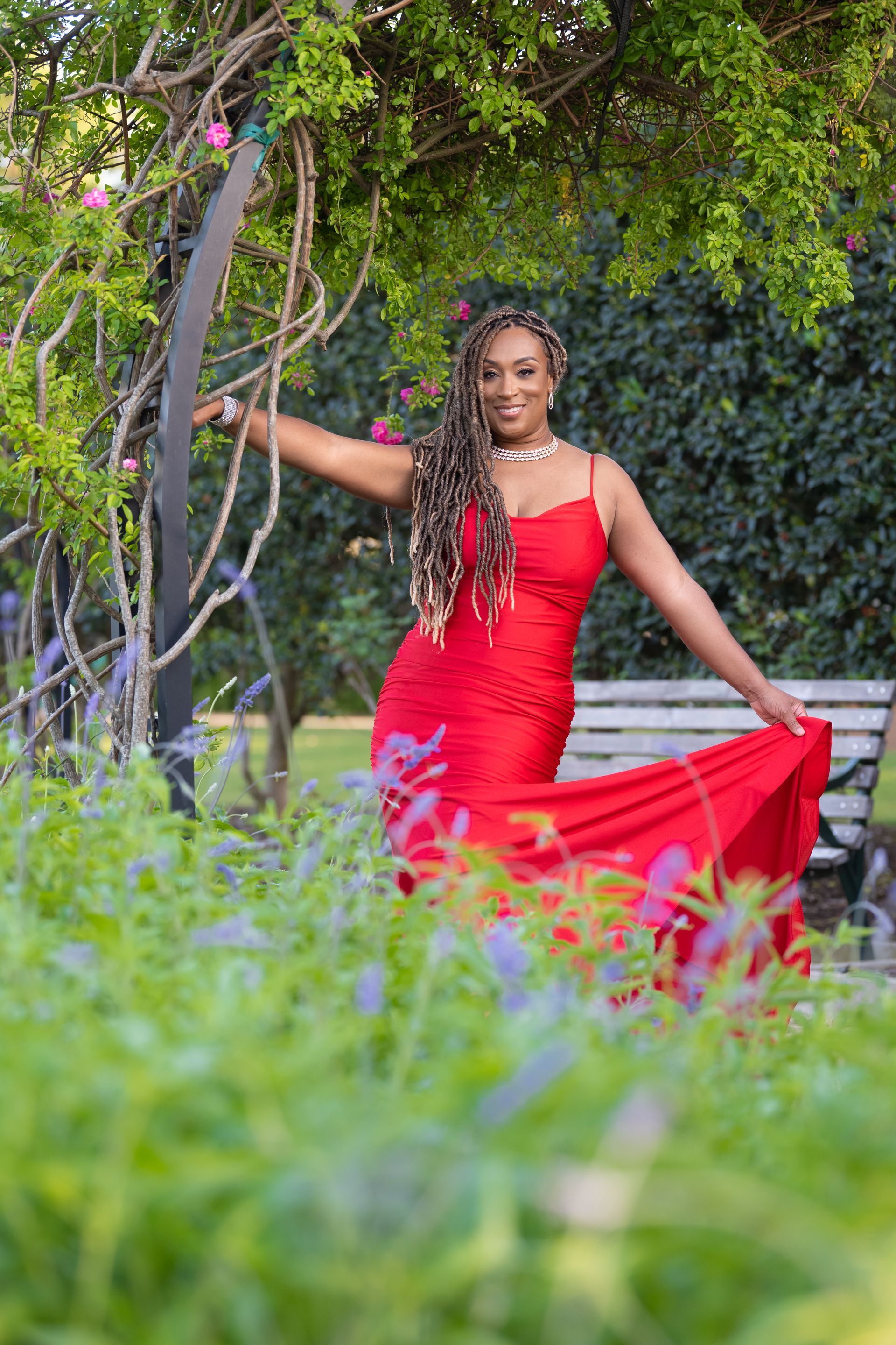 Woman in red dress poses outdoors with arm outstretched near green foliage.
