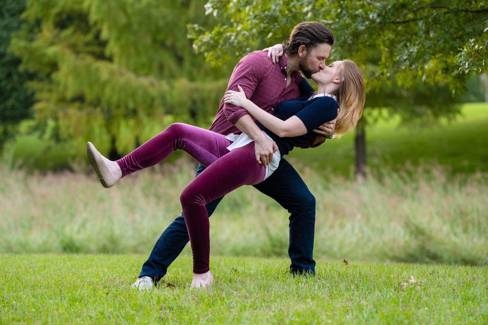Couple kissing in a grassy field; man holding woman who has one leg raised. Green trees in the background.