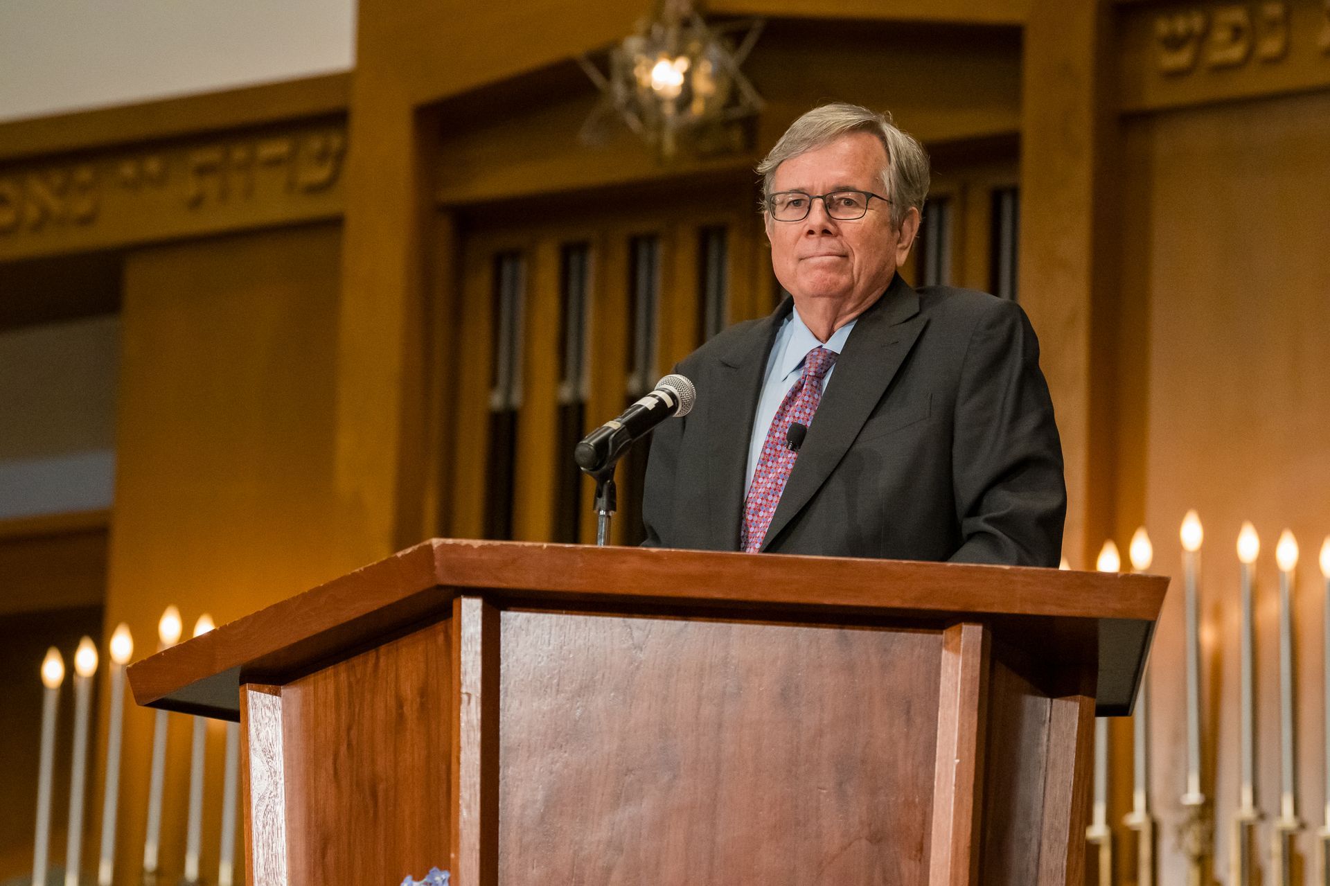 Man in suit speaking at a wooden podium in a synagogue. Candles and Hebrew text are visible.