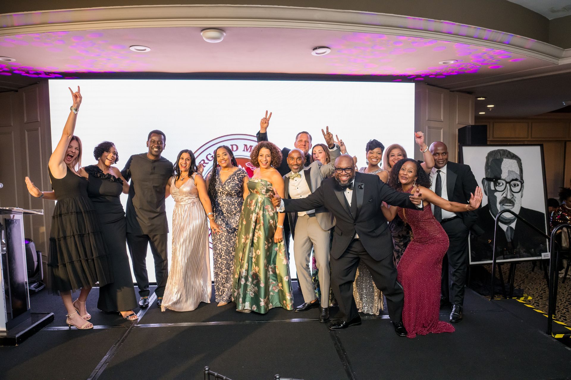Group of people on stage, celebrating.  Formal attire, raising arms, smiling. Backdrop with logo. Portrait on the right.