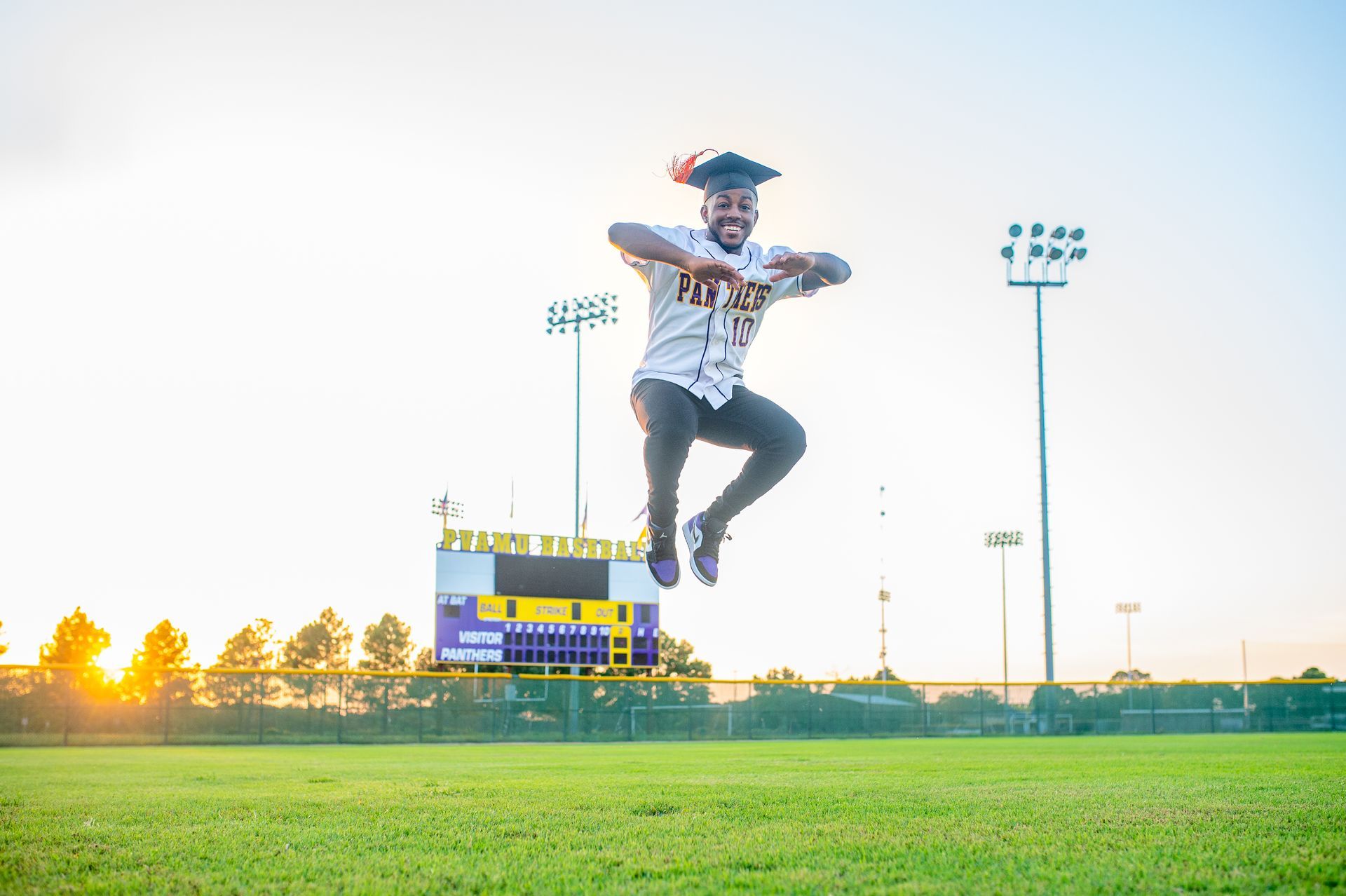 Man in graduation cap leaps for joy on a baseball field, scoreboard and sunset in background.