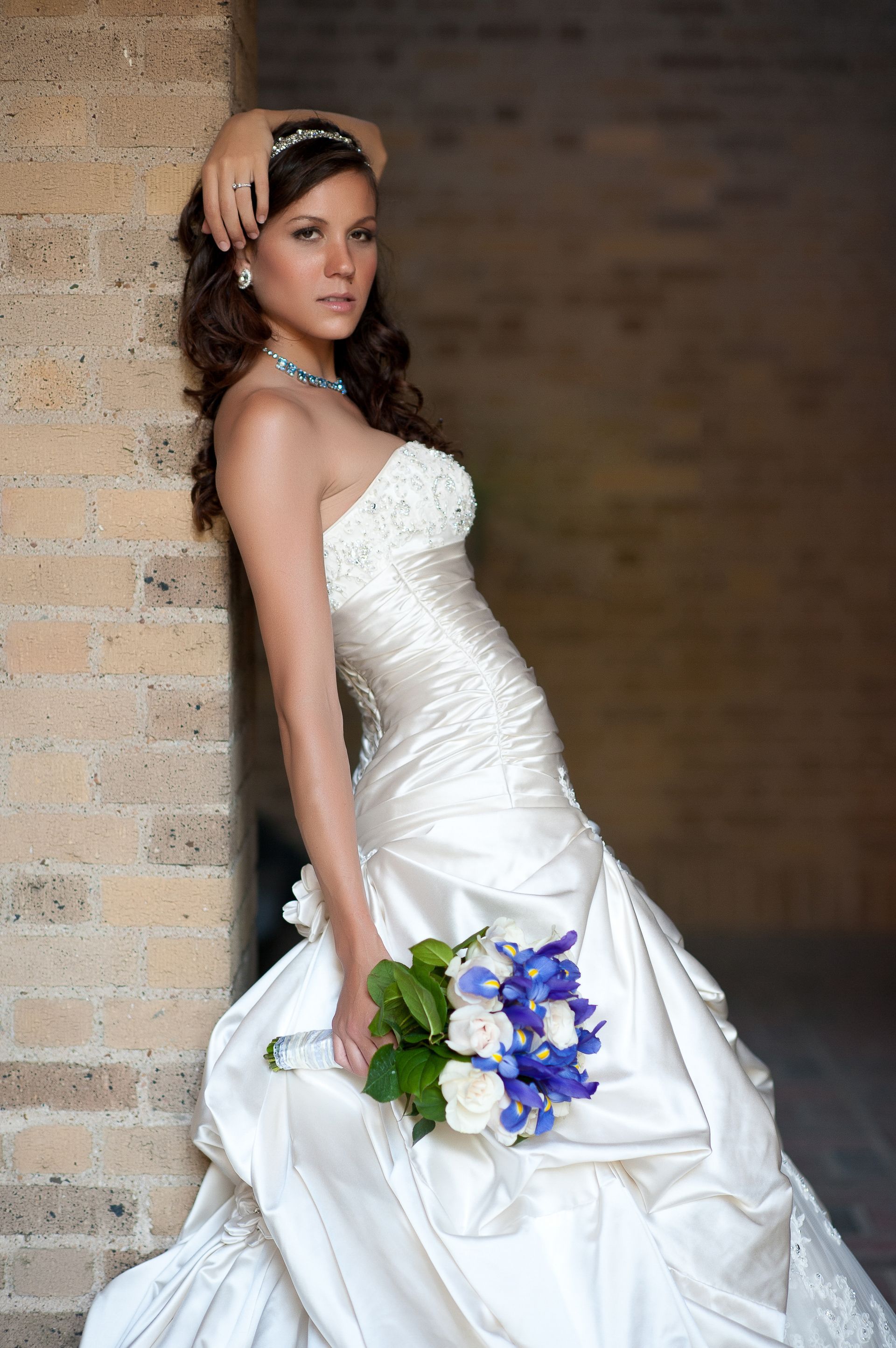 Bride in a white strapless gown, holding a bouquet, leaning against a brick wall.