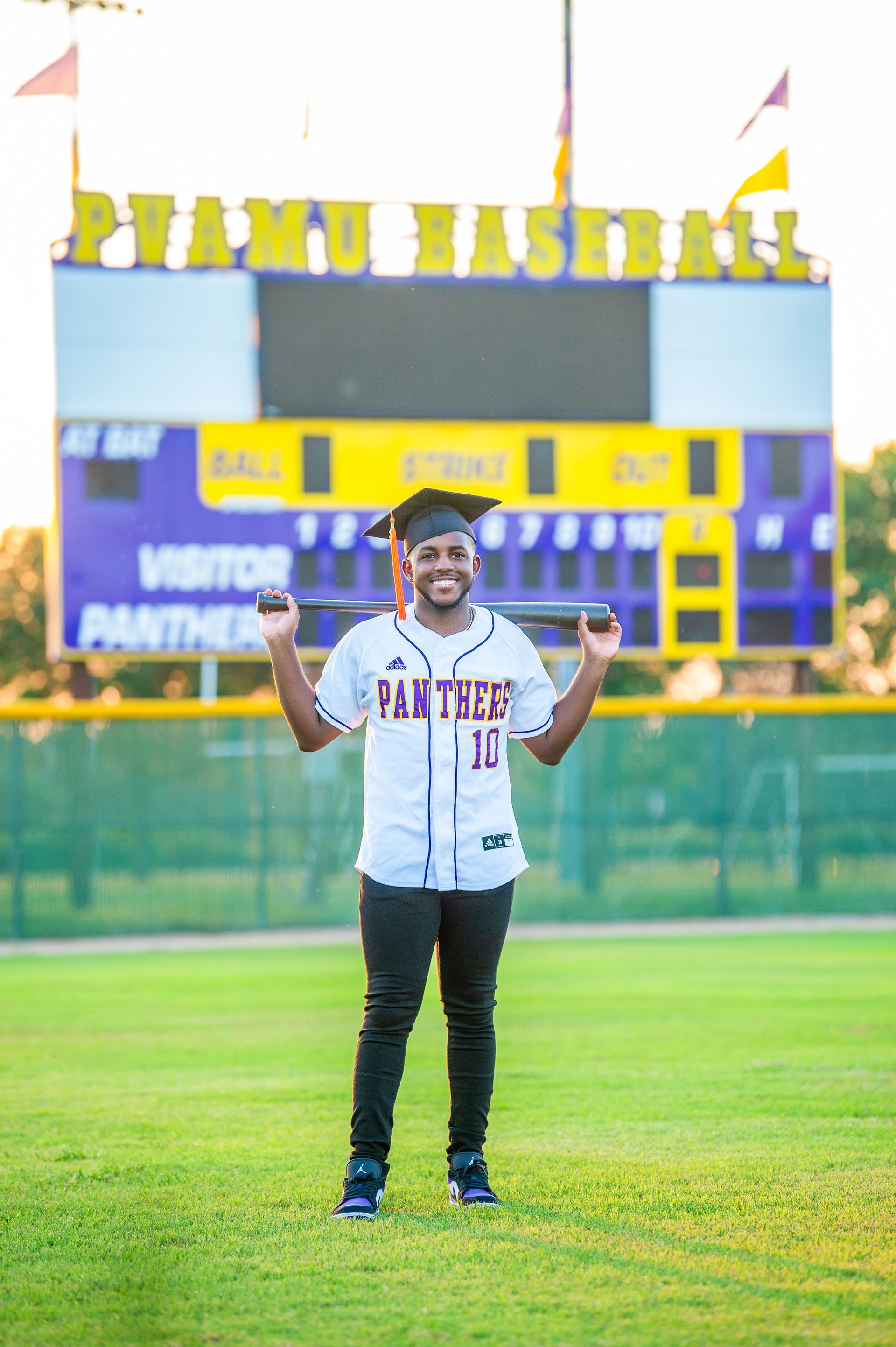Man in baseball jersey and graduation cap smiles, holding a bat on a baseball field with scoreboard.