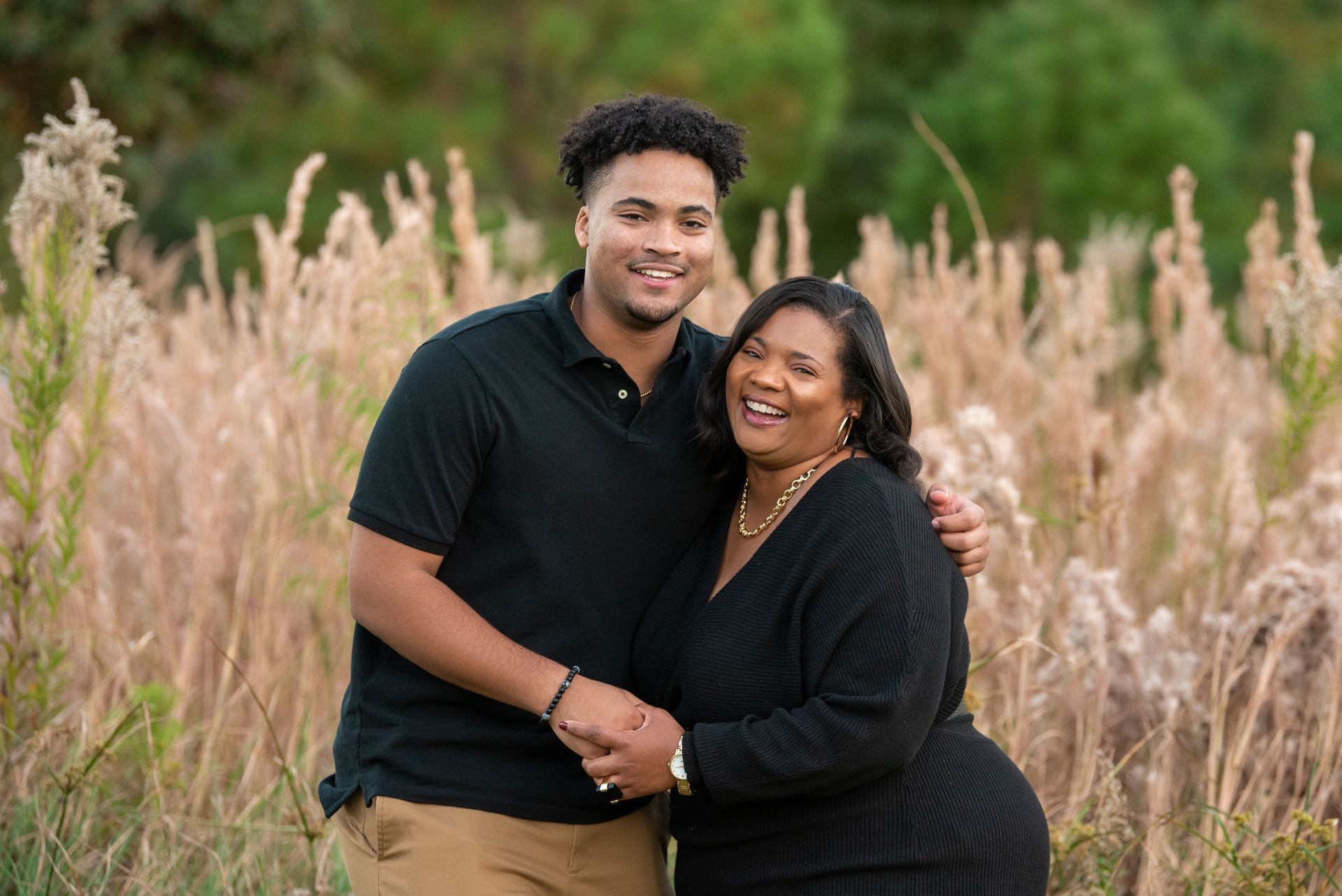 Two people smiling and embracing in a field of tall grass. Both are wearing black shirts.