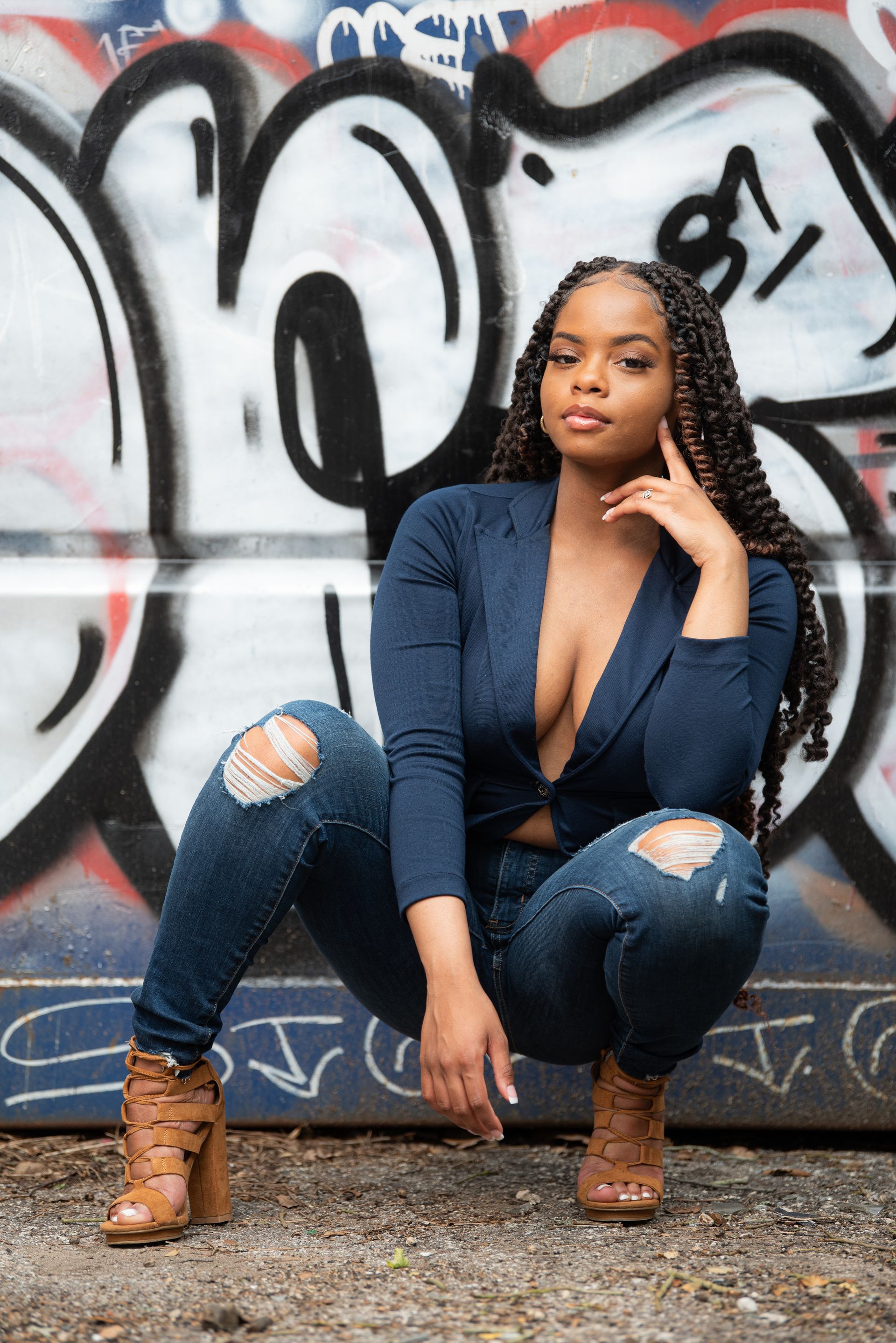 Woman in blue jacket and distressed jeans squats in front of graffiti.