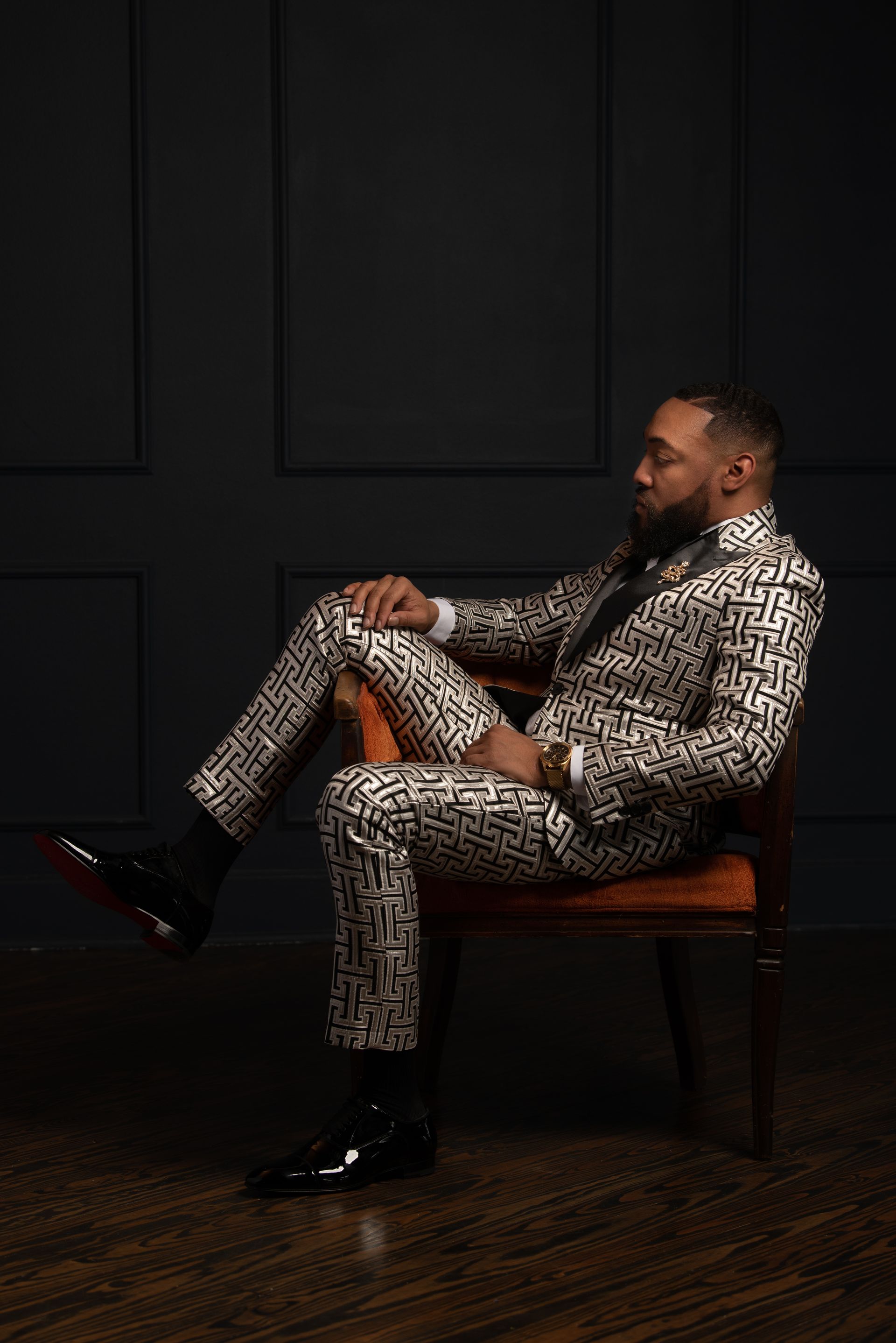 Man in patterned suit sits in an orange chair, black shoes, and dark background.