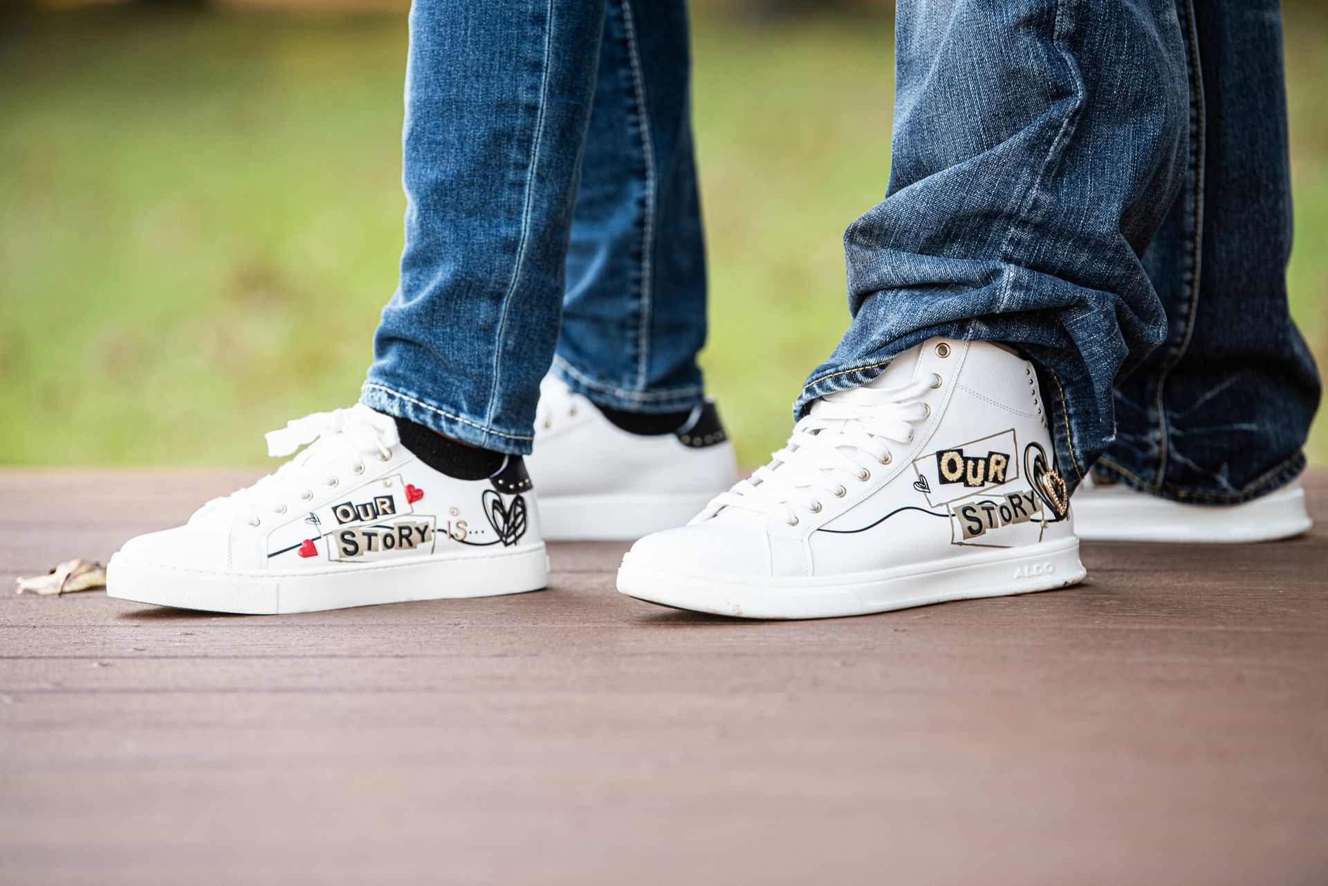 Two pairs of white sneakers with custom text and drawings, legs in blue jeans, standing on a wooden surface.
