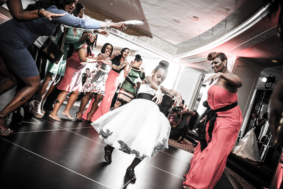 People dancing on a dance floor. One woman in white dress, another in red gown. Other guests watch.