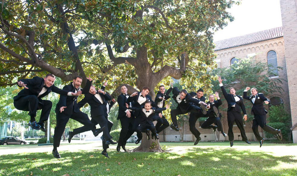 Groomsmen in black tuxedos jump in the air around a tree on a sunny day.