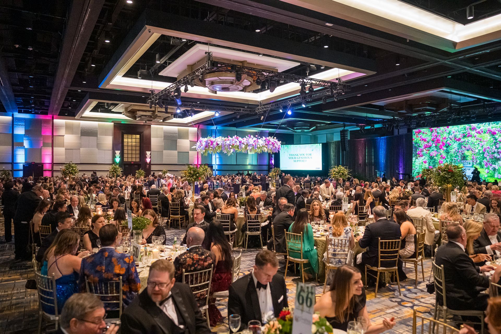 Large banquet hall filled with people at tables, many in formal attire. Bright lighting and floral arrangements.