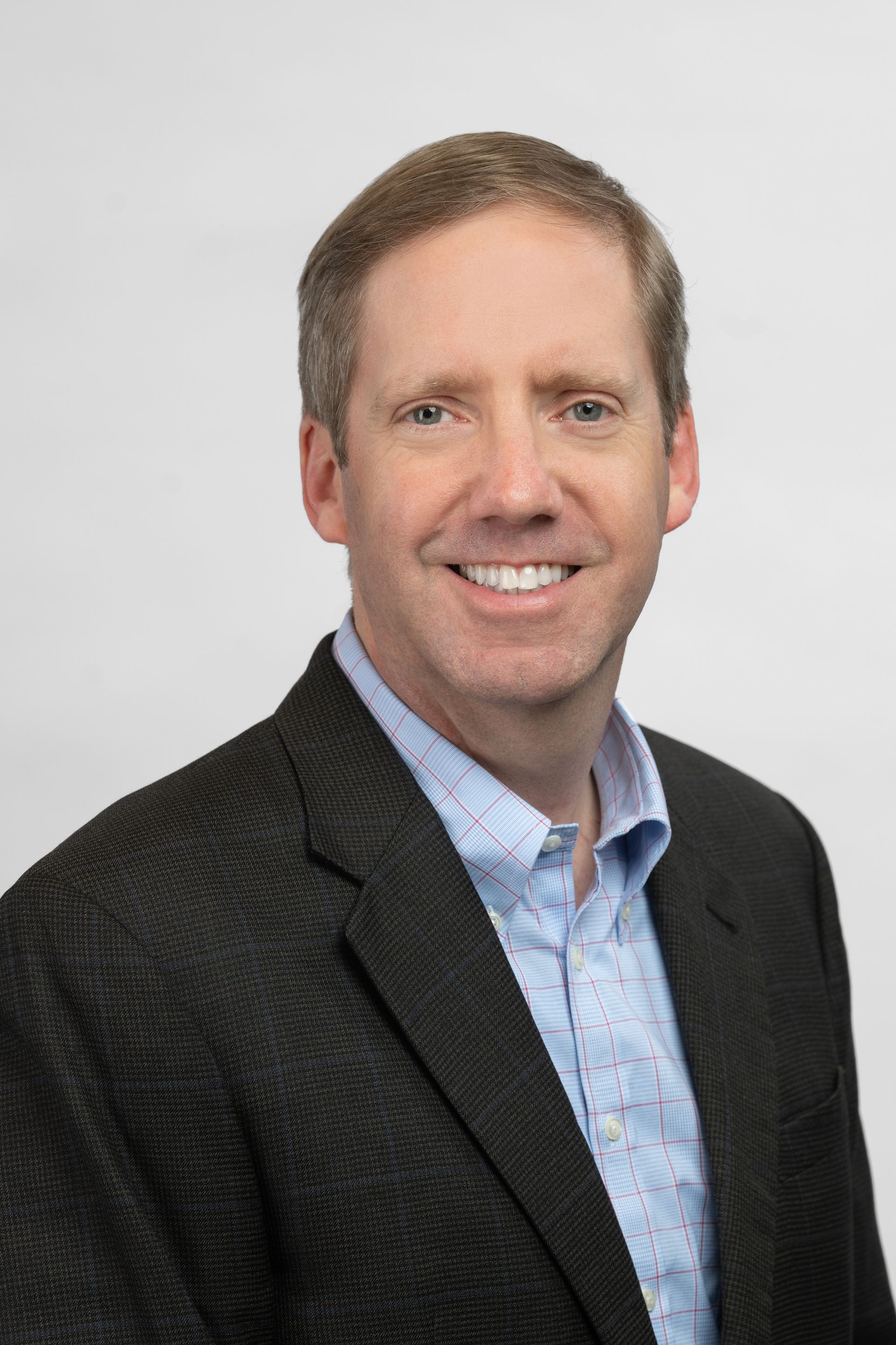 Man in a blazer and blue shirt smiles at the camera against a white backdrop.