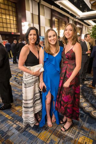 Three women in formal dresses at an event, posing. One in blue, one black and white, and one in floral.
