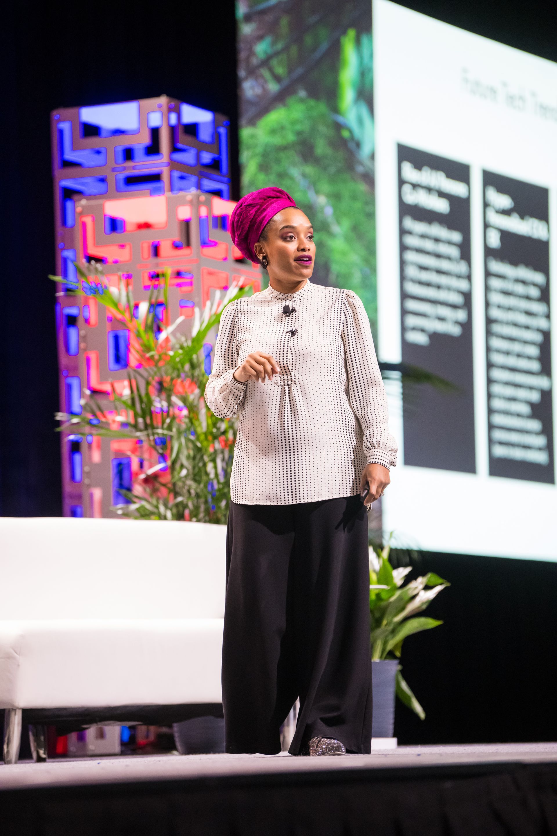 Woman in white patterned blouse, fuchsia headscarf, black pants, speaking on stage. Palm leaves, backdrop with text.