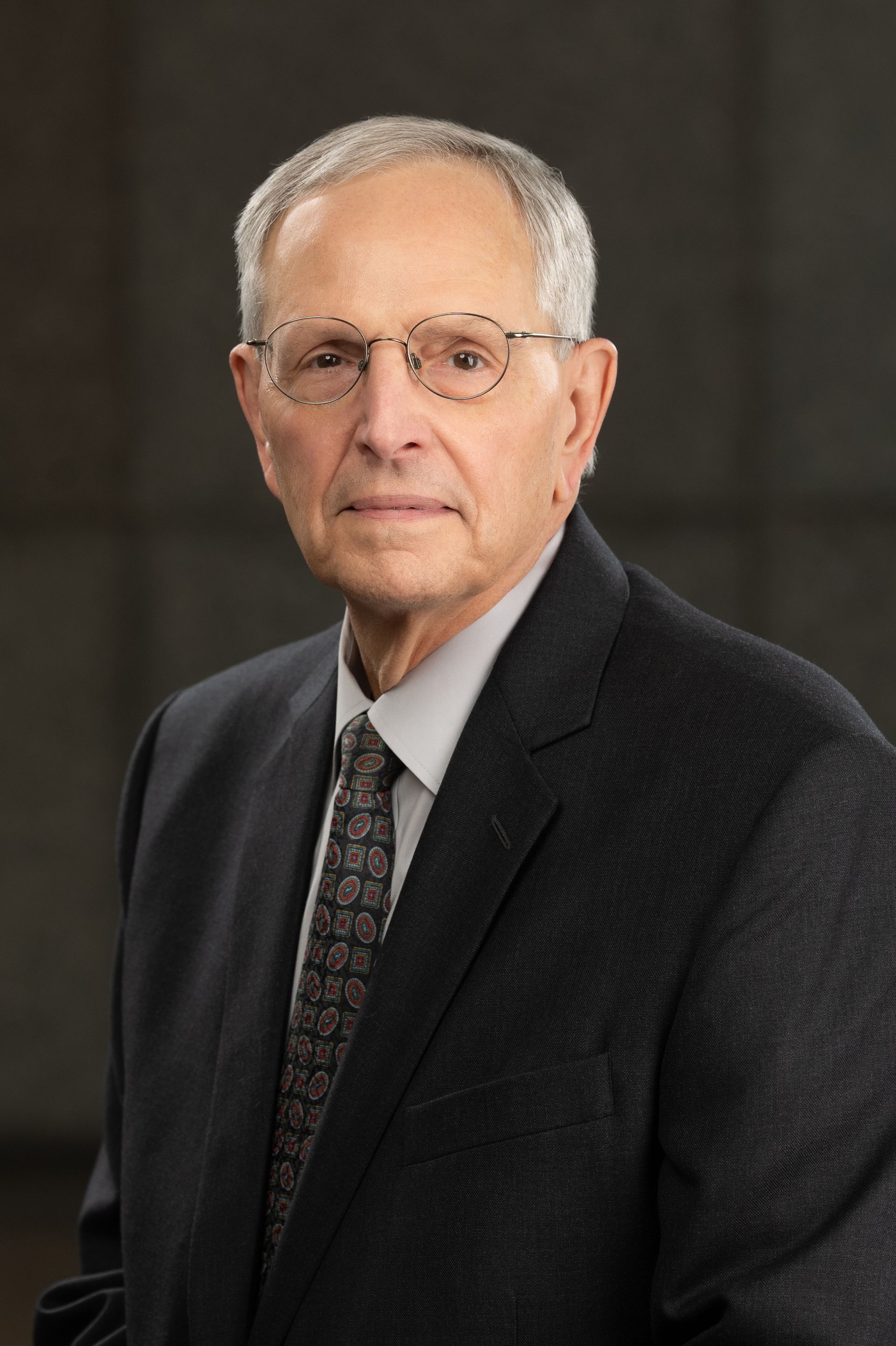 Man in glasses, suit, and tie, looking forward with a serious expression.