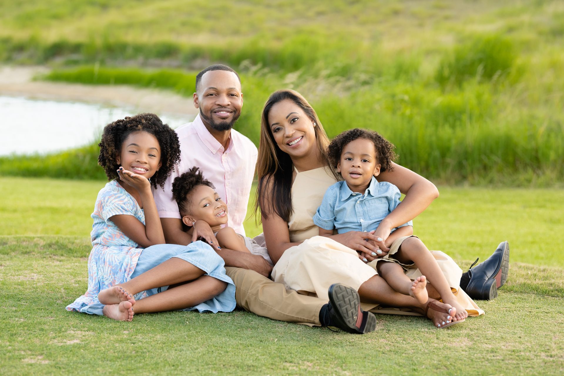 Family of five sitting on grass, smiling. Green background, sunny day.