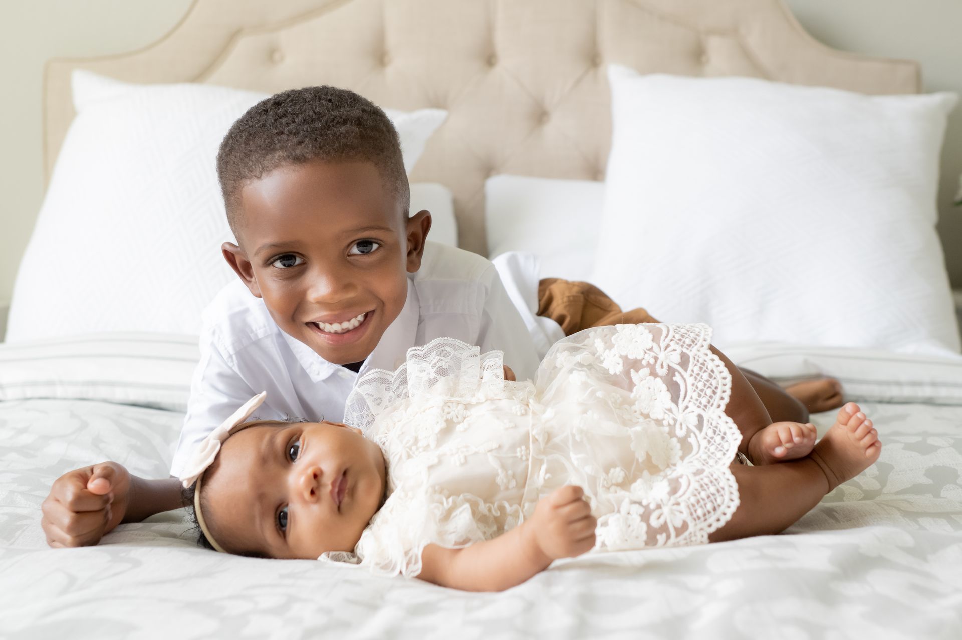 Boy smiles at camera, lying on a bed with a baby wearing a lacy dress and a dog, resting on the pillows.