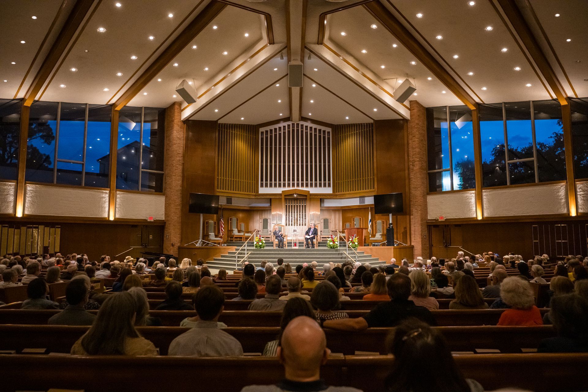 Audience in church observes speakers on stage. Brown wood interior, stained-glass windows, night sky visible.