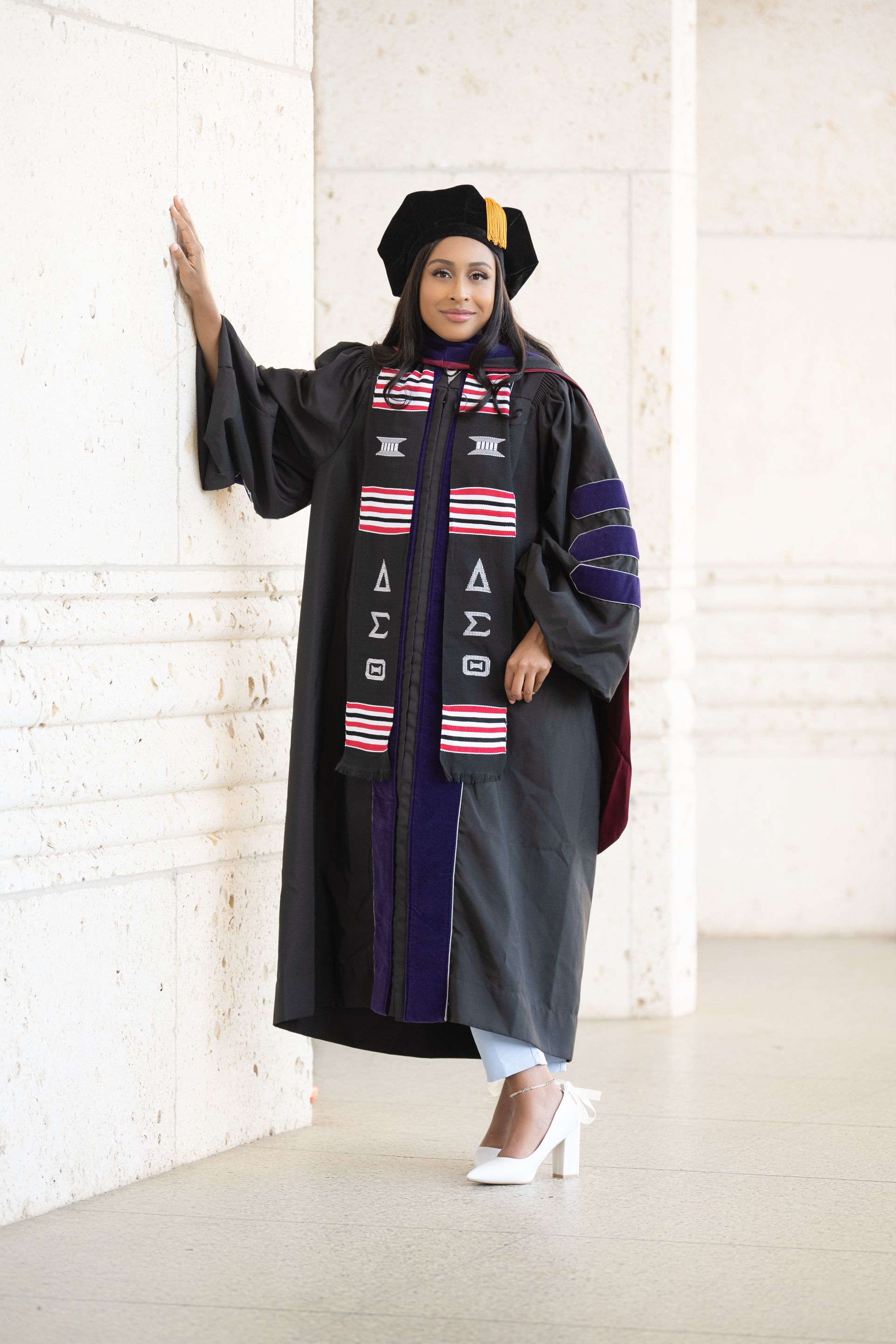 Person in graduation attire leaning against a wall, smiling. The gown is black with blue accents and a sash.