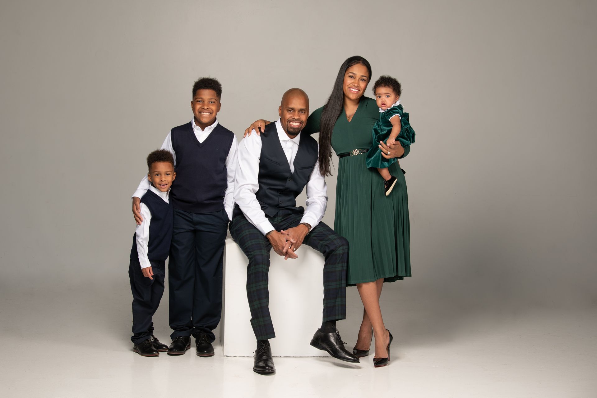 Family of five poses: Man seated on a white block, wife holding baby, and two sons stand near.