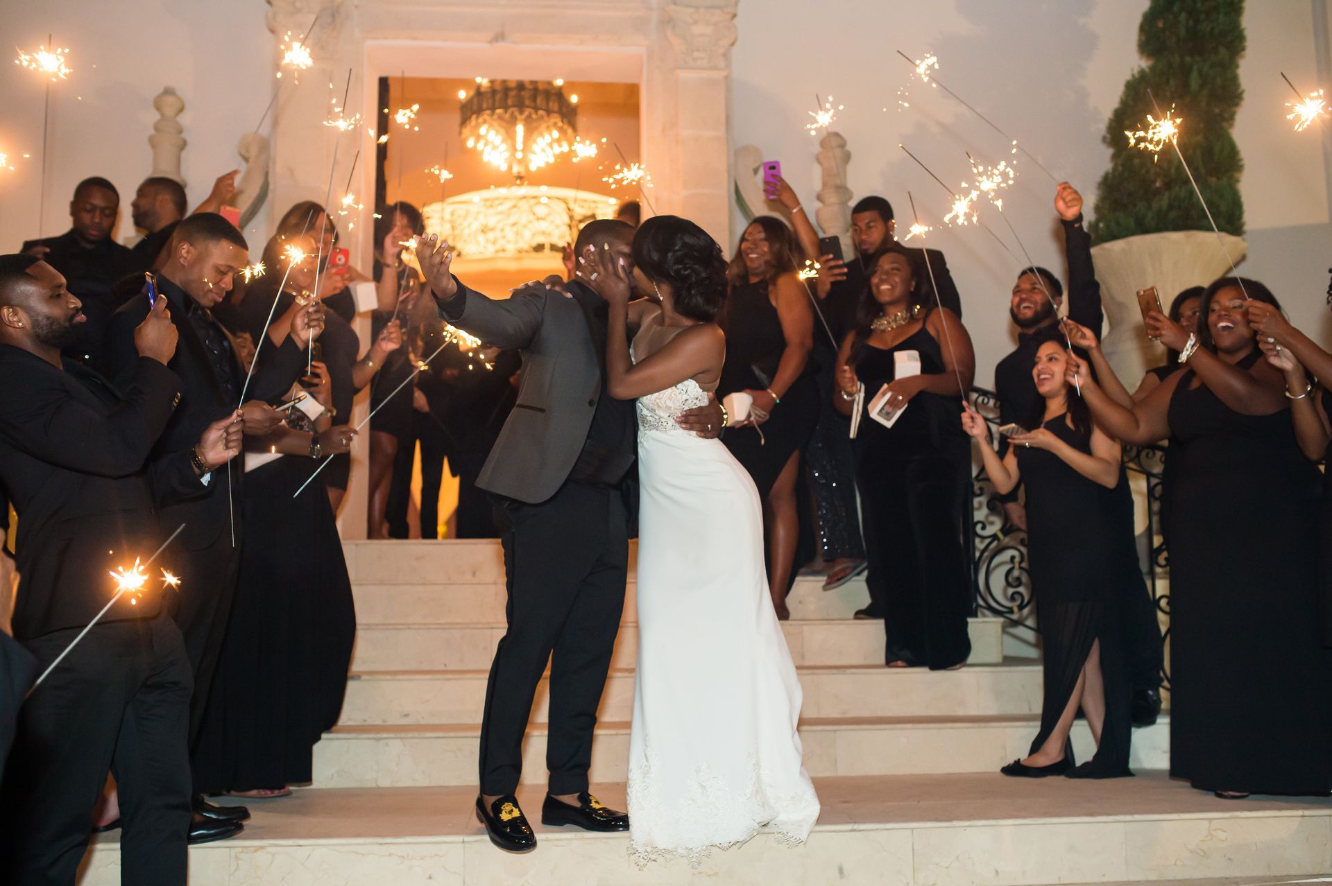 Bride and groom kiss on steps as guests hold sparklers.
