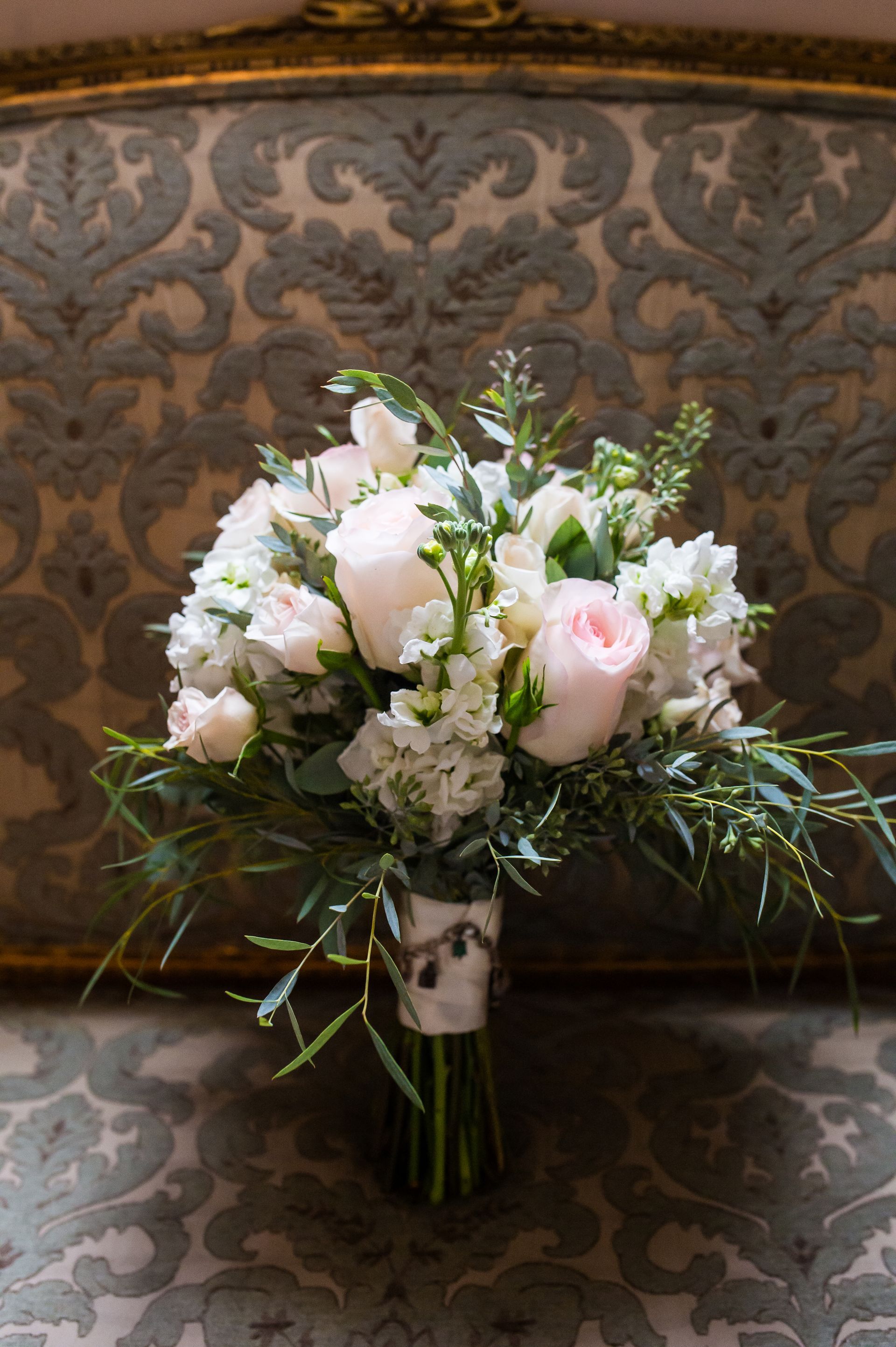 Wedding bouquet of pink roses and white flowers, wrapped in ribbon, on ornate patterned upholstery.
