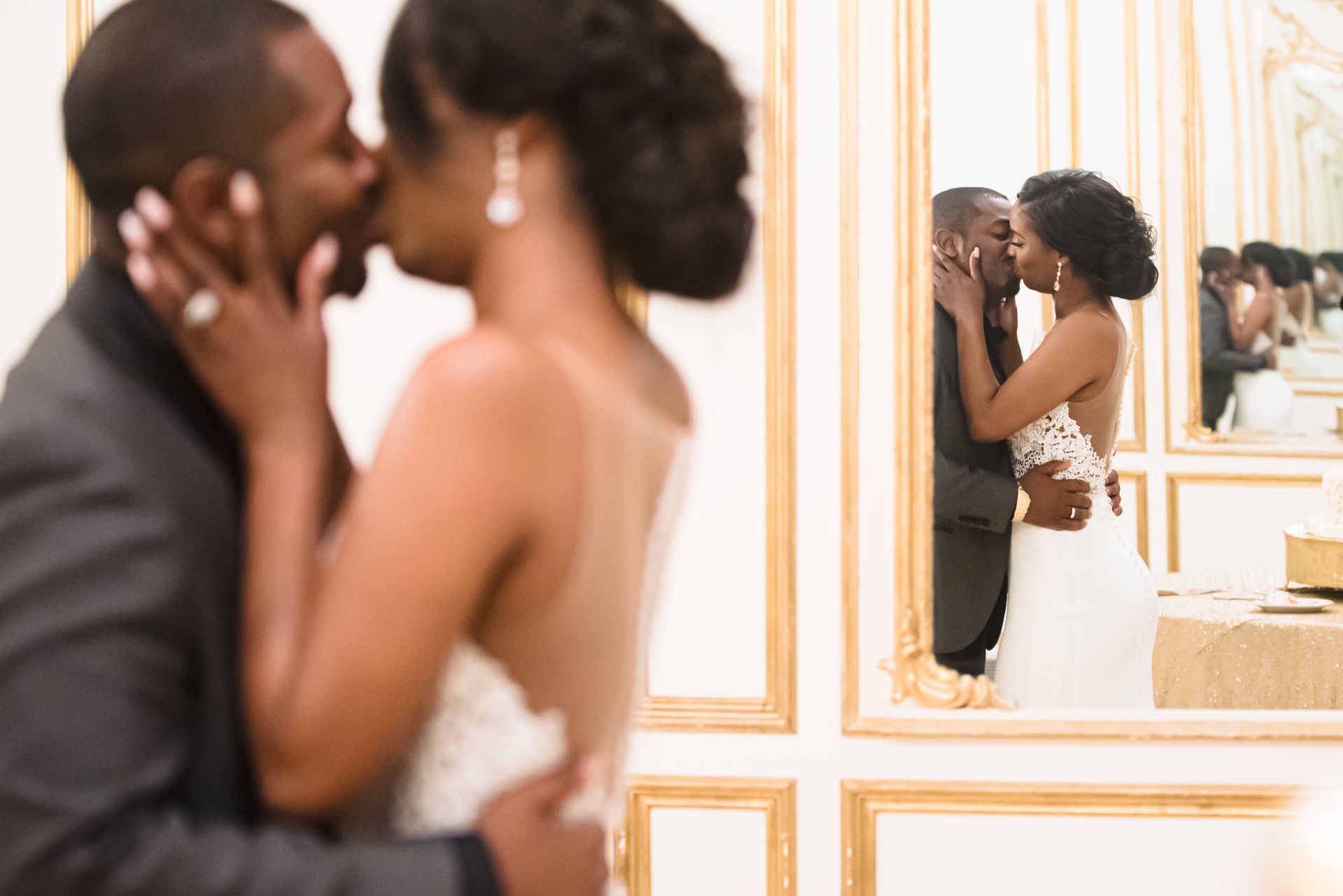 Couple kissing in front of a mirror, bride in white dress, groom in gray suit, gold-framed mirror reflects the kiss.