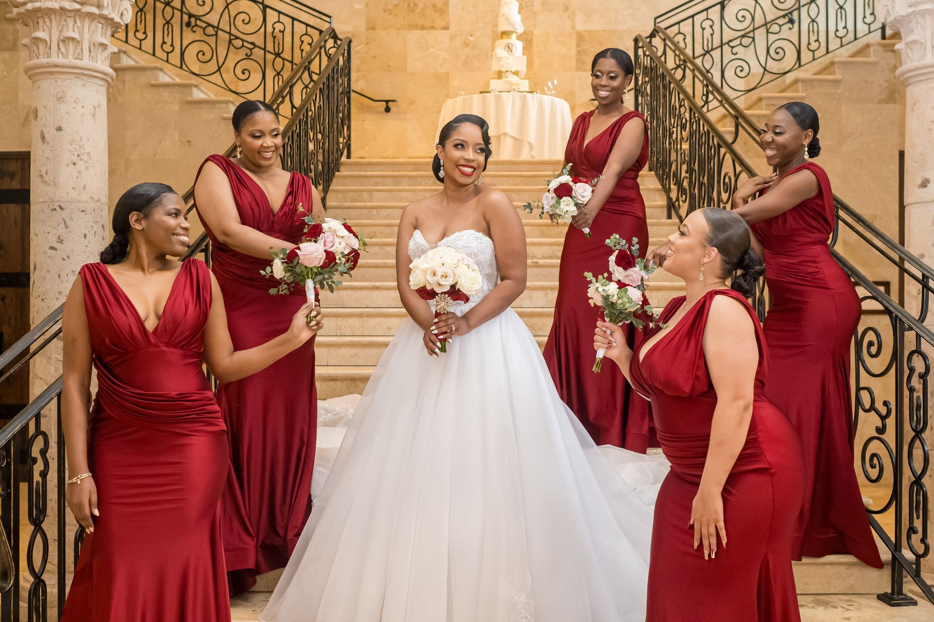 Bride in white gown surrounded by bridesmaids in red dresses on a staircase, smiling.