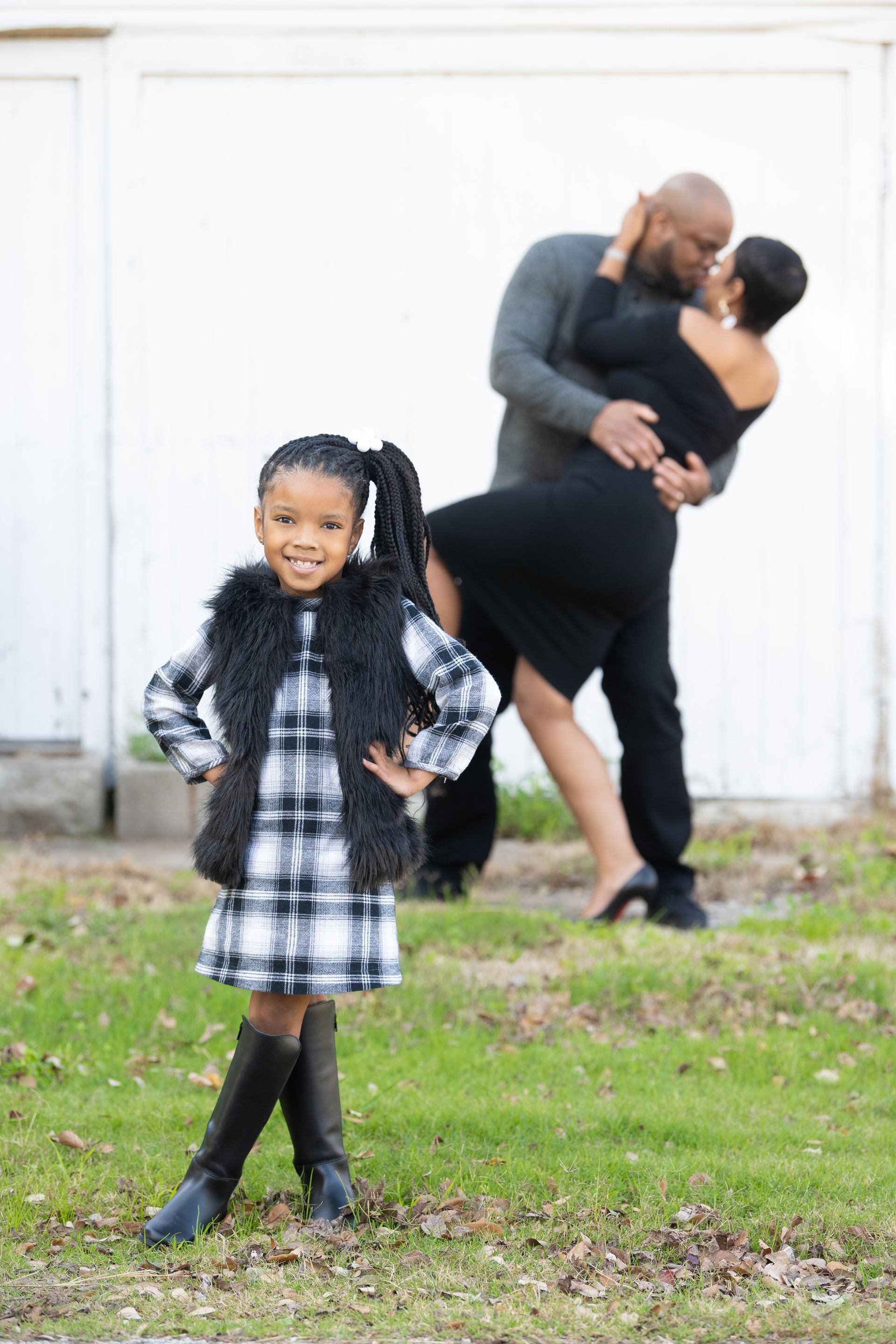 Girl in plaid dress and vest poses, parents dance and embrace in background.