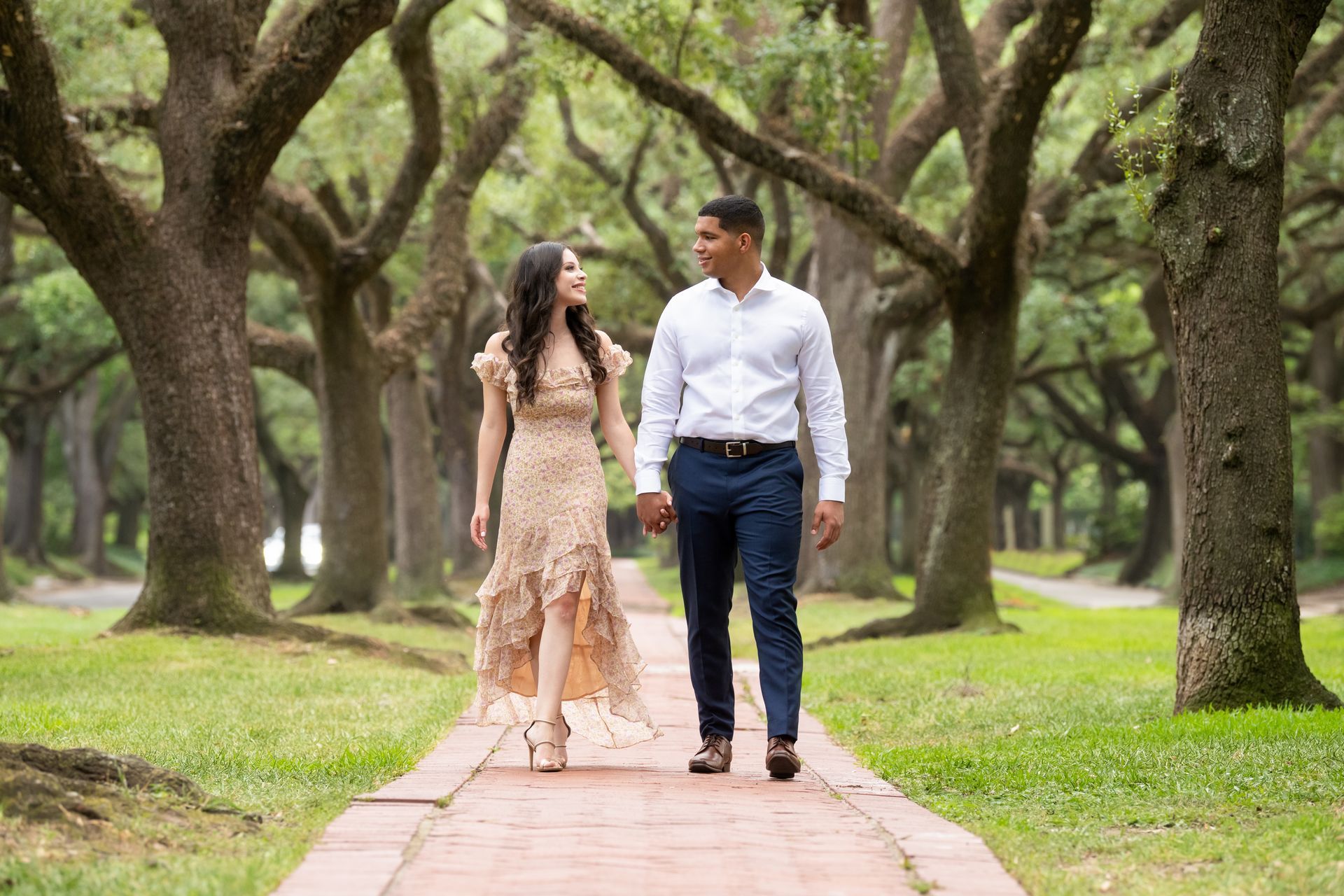 Couple walking hand-in-hand on brick path under tree-lined canopy. They are smiling and looking at each other.
