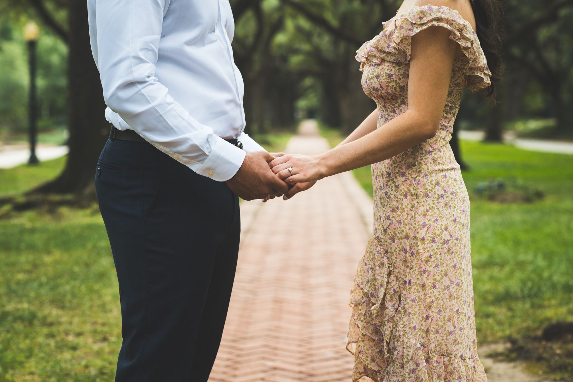 Couple holding hands, standing on a brick path, trees in background.