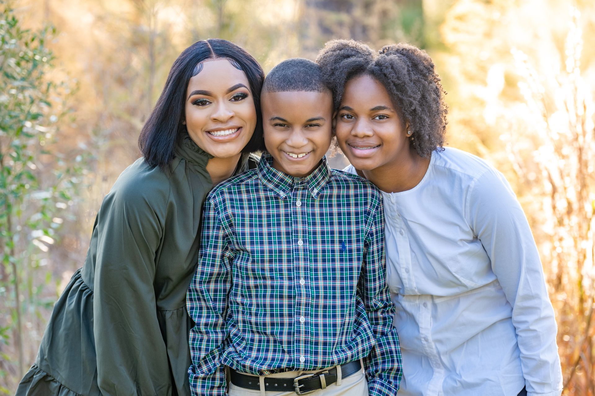 Three people smiling, posed outdoors. One person wears a green dress, one a plaid shirt, and one a white shirt.