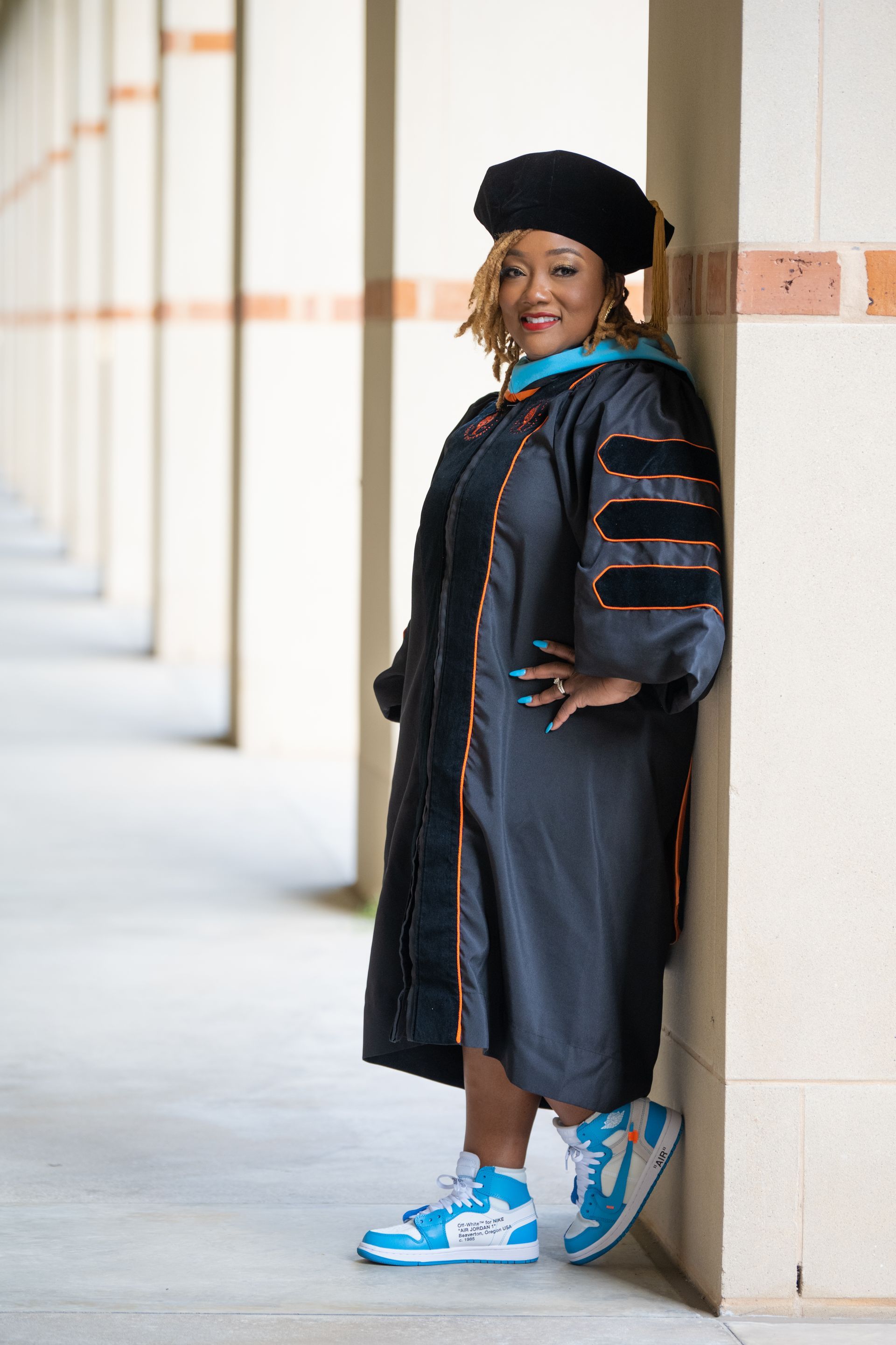 Person in graduation gown and cap leans against a building's pillars, wearing blue and white sneakers; smiles.