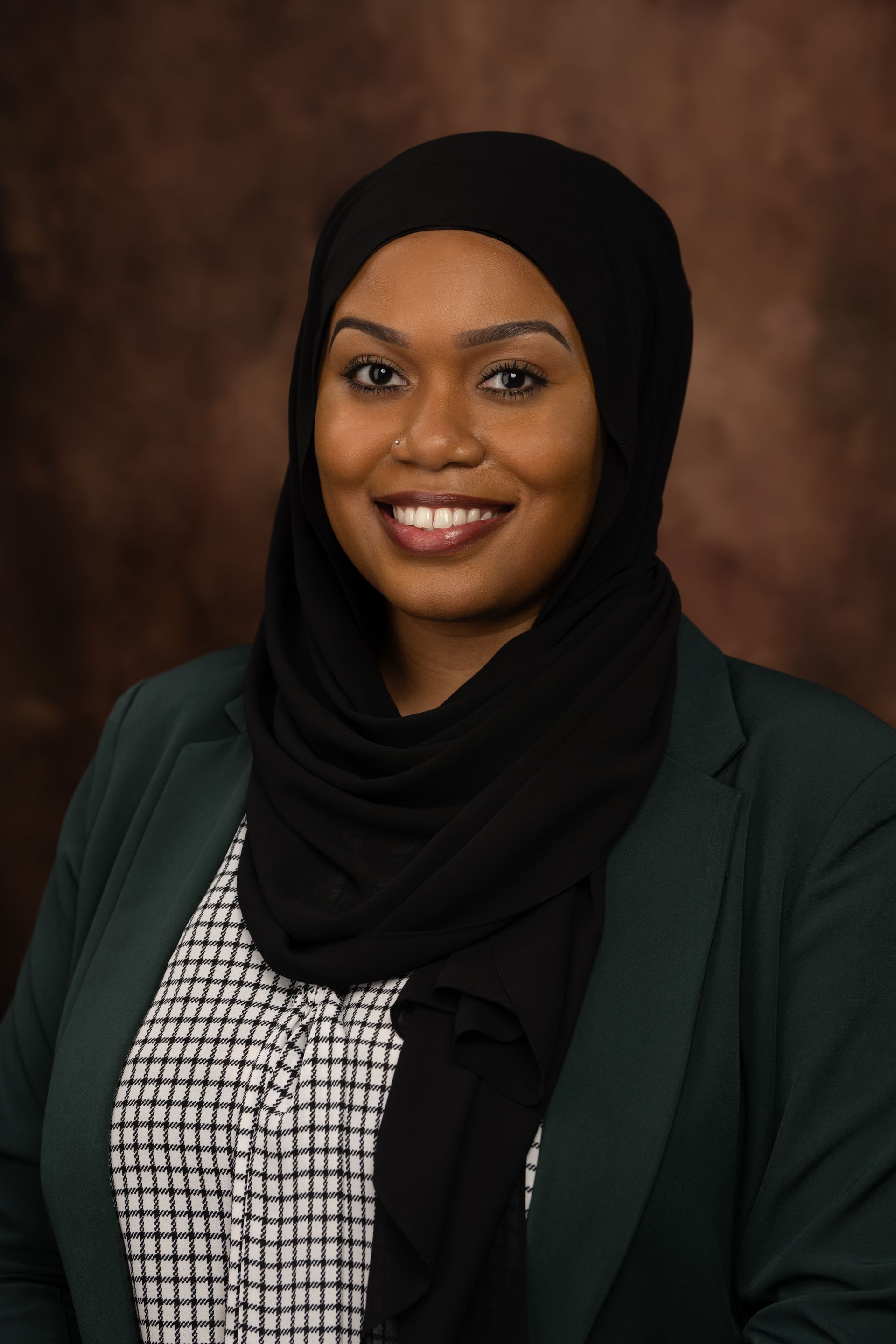 Woman in black hijab and green jacket smiling at the camera, against a brown backdrop.