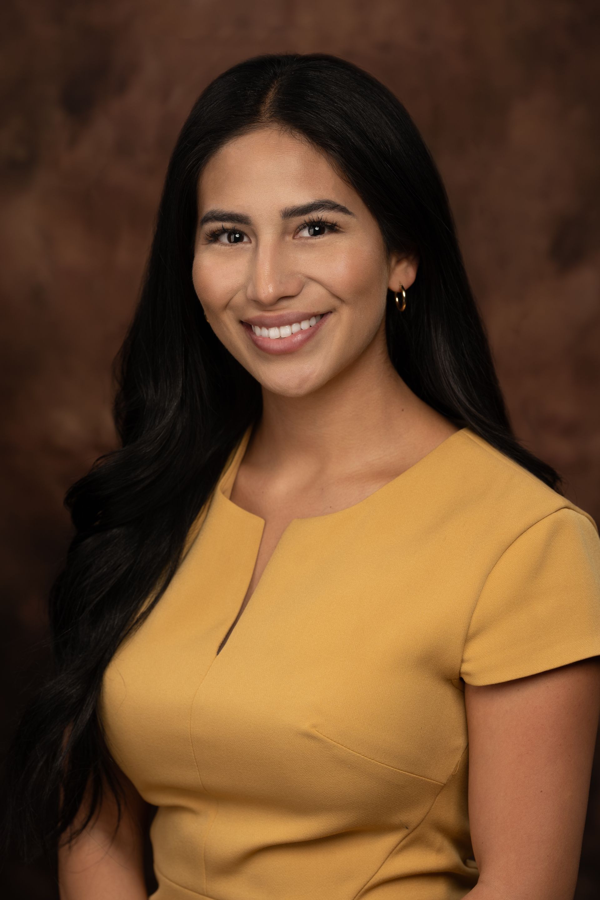 Woman with long dark hair smiles in a yellow dress; brown backdrop.